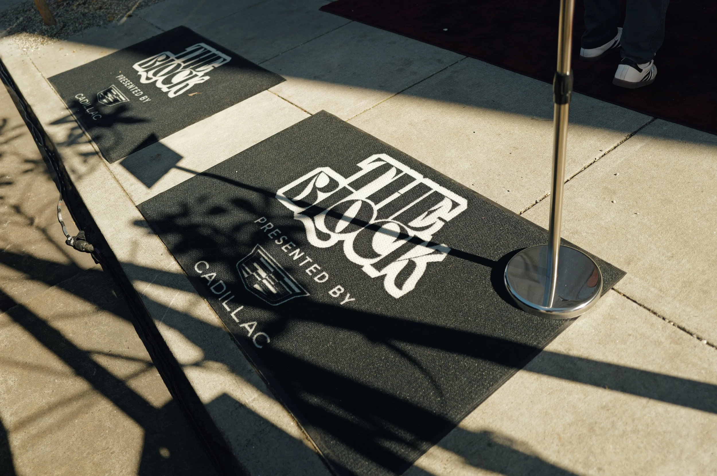 Two black mats with white text and logos on a sidewalk, with shadows and part of a standee or pole visible.