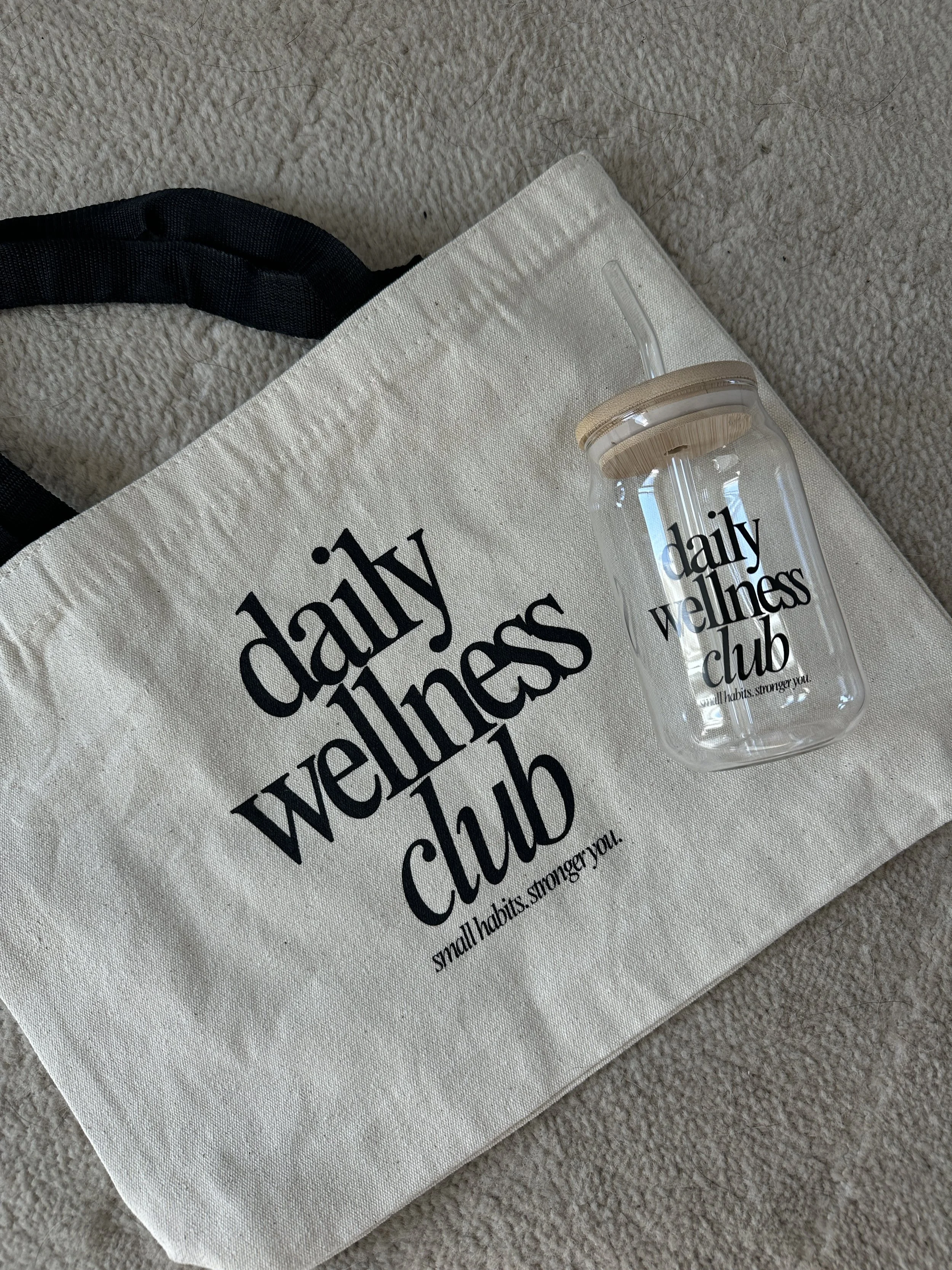 A white tote bag with black text that reads 'daily wellness club' and a glass bottle with a wooden lid and straw, placed on a beige carpet.