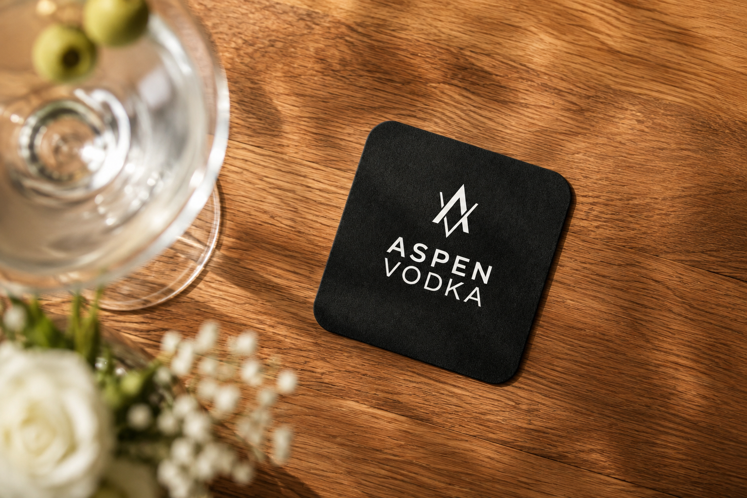 A black coaster with a white logo and text that reads 'Aspen Vodka' on a wooden table, with a glass and white flowers partially visible in the foreground.