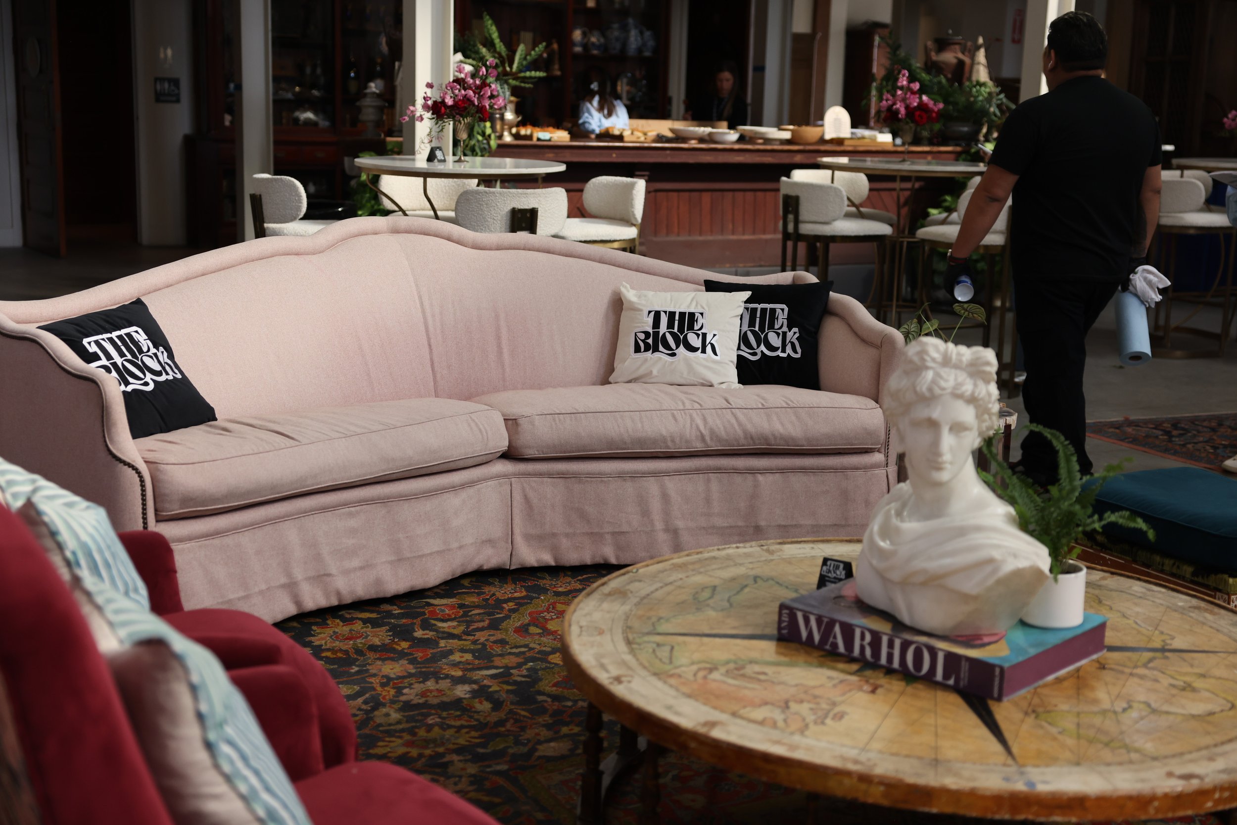 A cozy living room or waiting area with a pink sofa adorned with black and white pillows that read "The Block," a wooden round coffee table with books and a white sculpture, and a variety of other chairs and tables with floral arrangements. There are