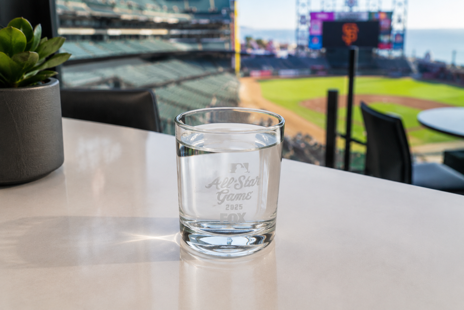 A glass of water on a white table with a baseball field in the background, seen from a stadium or sports club with seating and a large scoreboard.