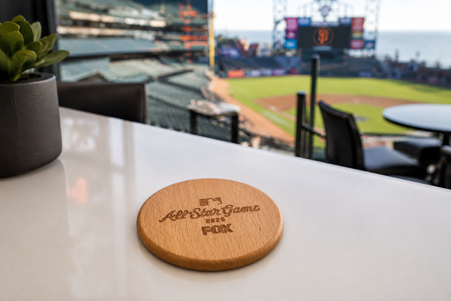 Wooden coaster engraved with "All-Star Game 2025 FOX" on a white table in a suite at a baseball stadium, with view of the field and stands in the background.