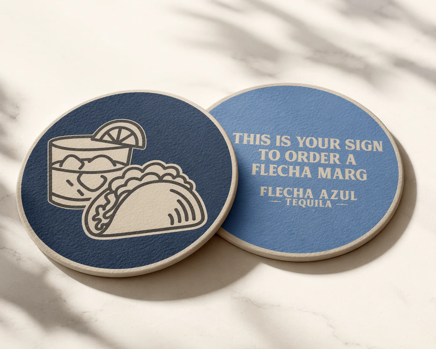 Two round signs on a white surface. The first sign shows a line drawing of a glass of a margarita and a taco. The second sign reads, "This is your sign to order a Flecha Marg, Flecha Azul Tequila."