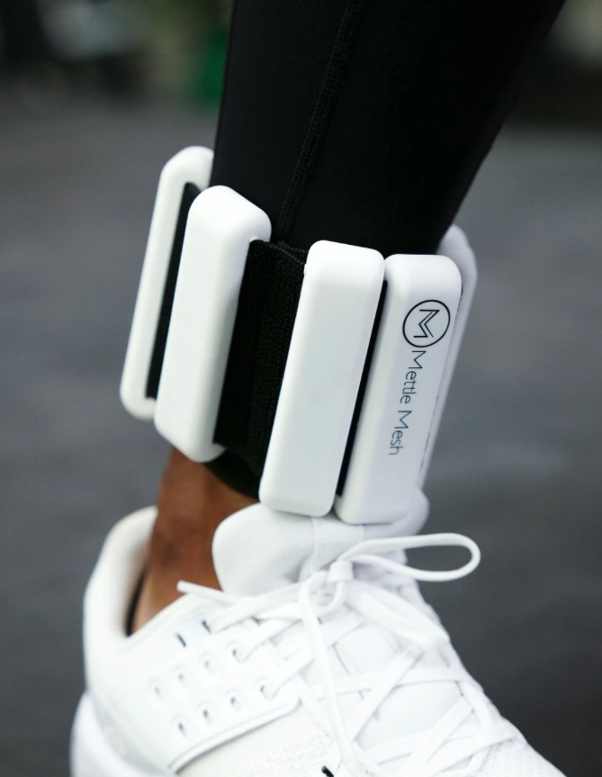 Close-up of an ankle with a white Metta Mesh wearable device secured with black straps over a white sneaker and black pants.