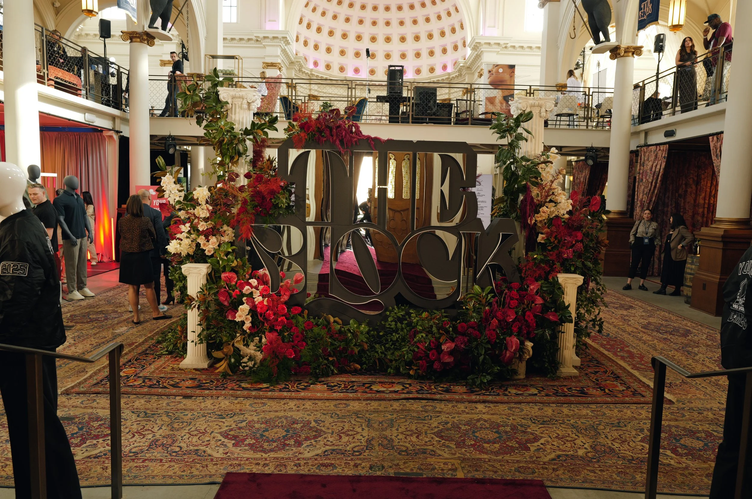 Indoor event space decorated with flowers and a large love sign for a wedding or celebration. There are people walking and standing, some on a mezzanine level, with ornate architecture and draped curtains. The ceiling is dome-shaped with gold accents