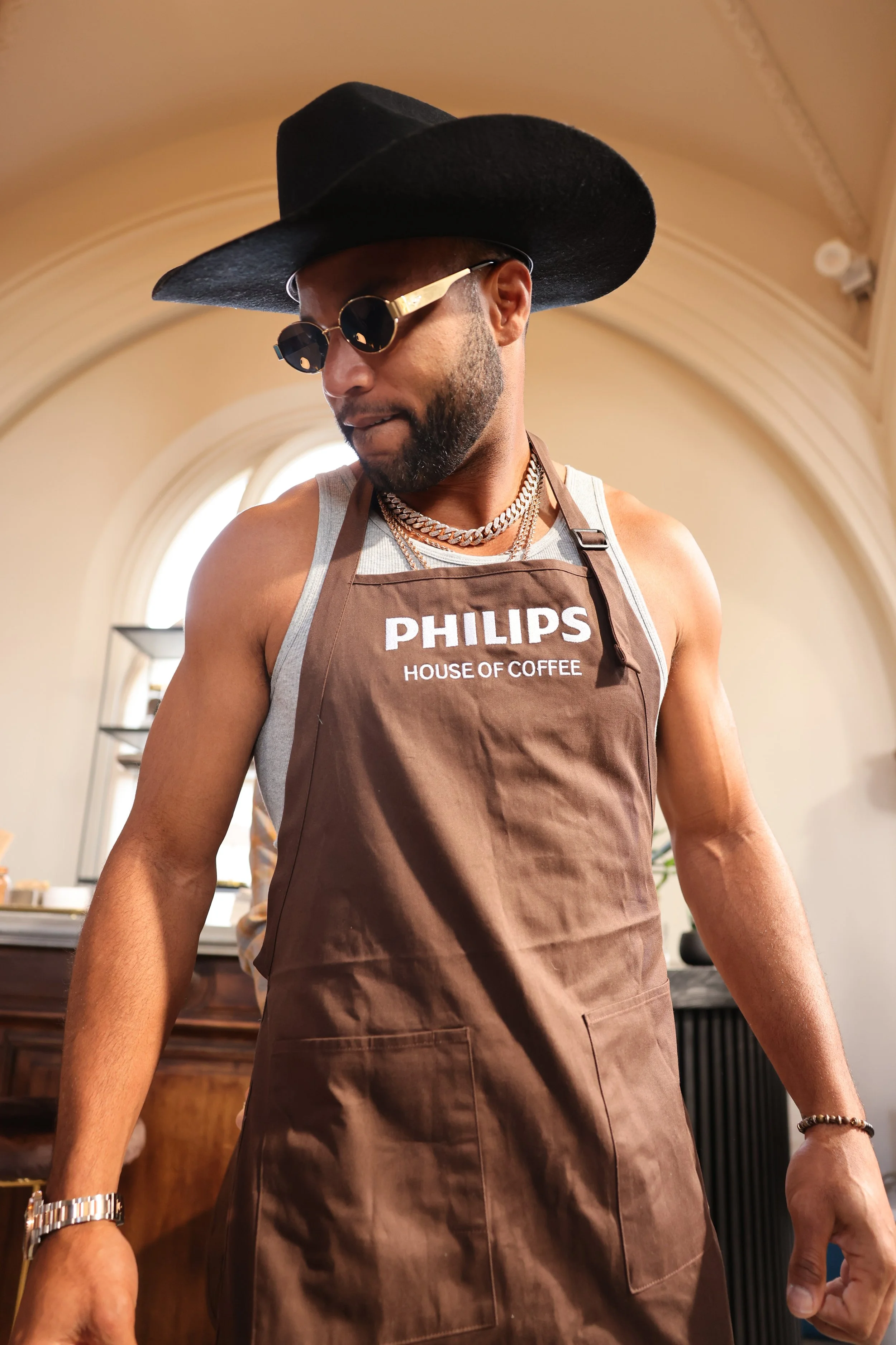 A man wearing a large black wide-brimmed hat, sunglasses, a sleeveless shirt, a brown apron with 'Philips House of Coffee' written on it, and multiple silver chains, standing indoors with an arched window and decorative molding in the background.