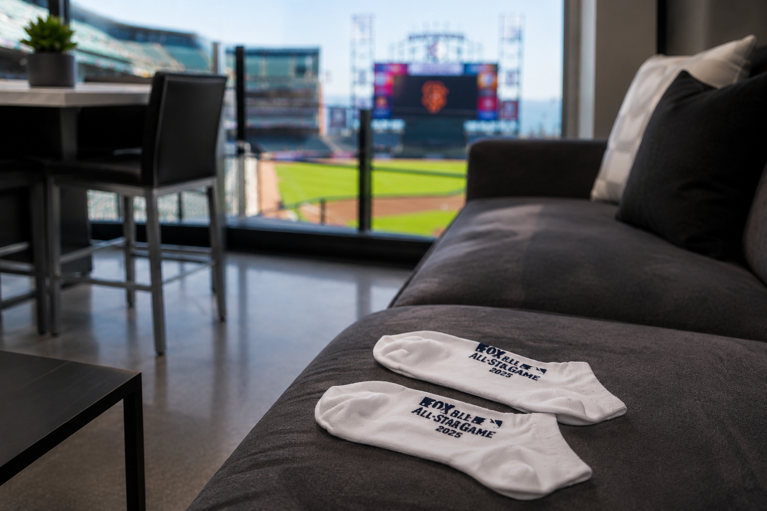 Two white socks with blue text and logo reading 'FOX VALLEY ALL-STAR GAME 2025' lying on a dark gray sofa in a living room with a baseball stadium view through large windows.