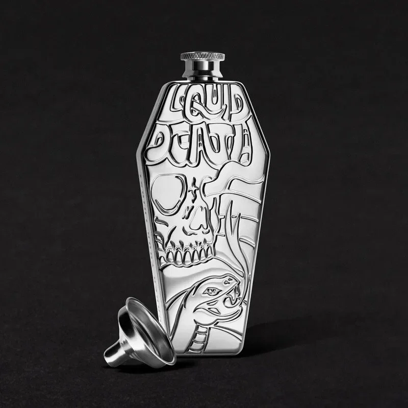 Silver flask with embossed skull, snake, and text reading 'Good Death,' placed against a black background, with a small funnel beside it.