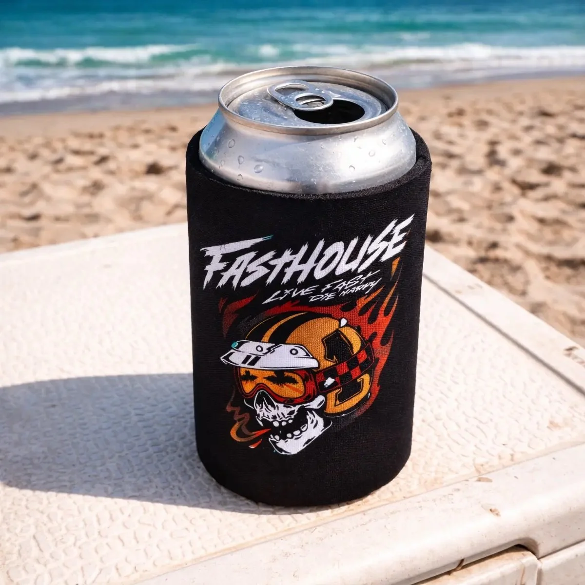 A can of beer in a black koozie with a graphic of a skull wearing a helmet and goggles, with the text 'Fast House Live Fast Ride Free,' placed on a white surface at the beach with sand and the ocean in the background.