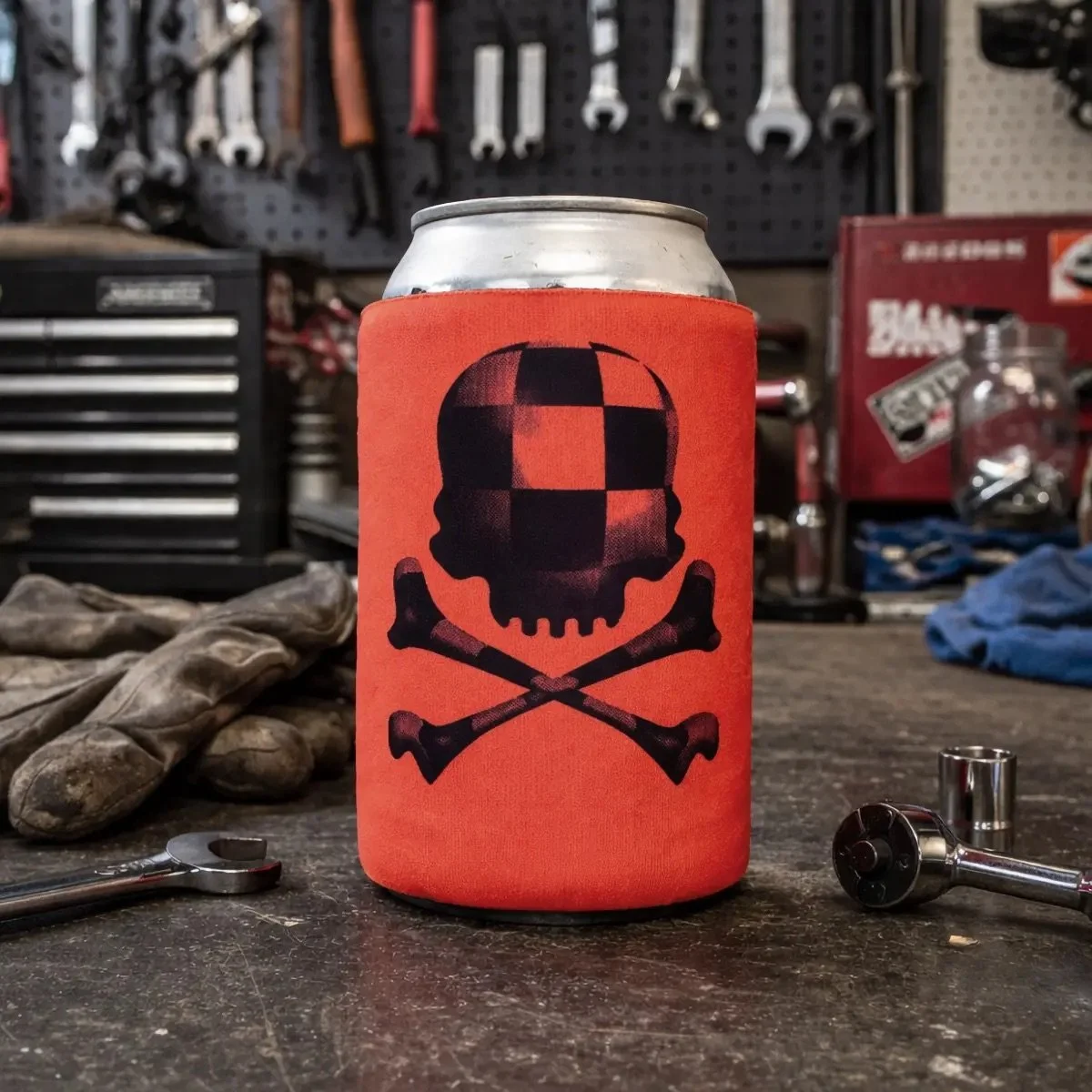 A can with a black skull and crossbones design on an orange background, placed on a workshop table with tools and gloves nearby.