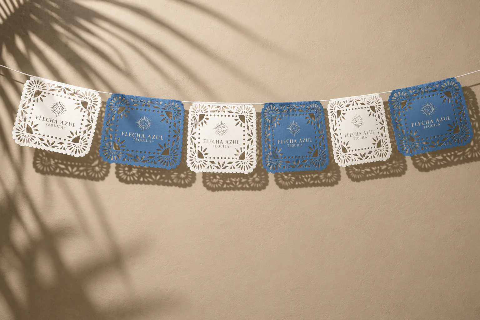Decorative paper banners with a cut-out floral pattern, alternating between white and blue, hanging against a beige wall with shadows of a plant's leaves.