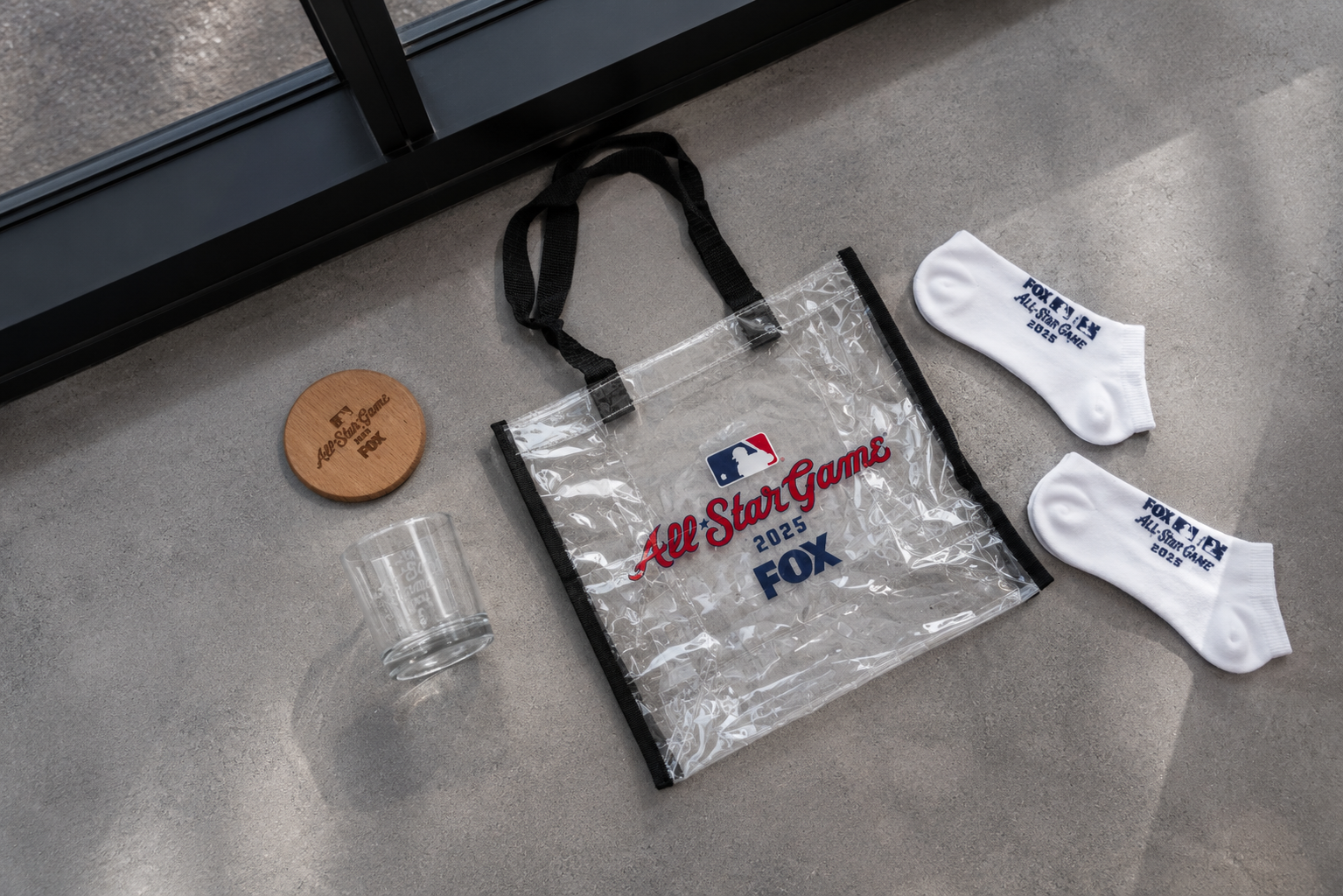 MLB All-Star Game 2025 FOX tote bag, two white socks with blue text, a clear glass, a wooden circular item with text, and a clear plastic cup all placed on a gray surface near a glass door.