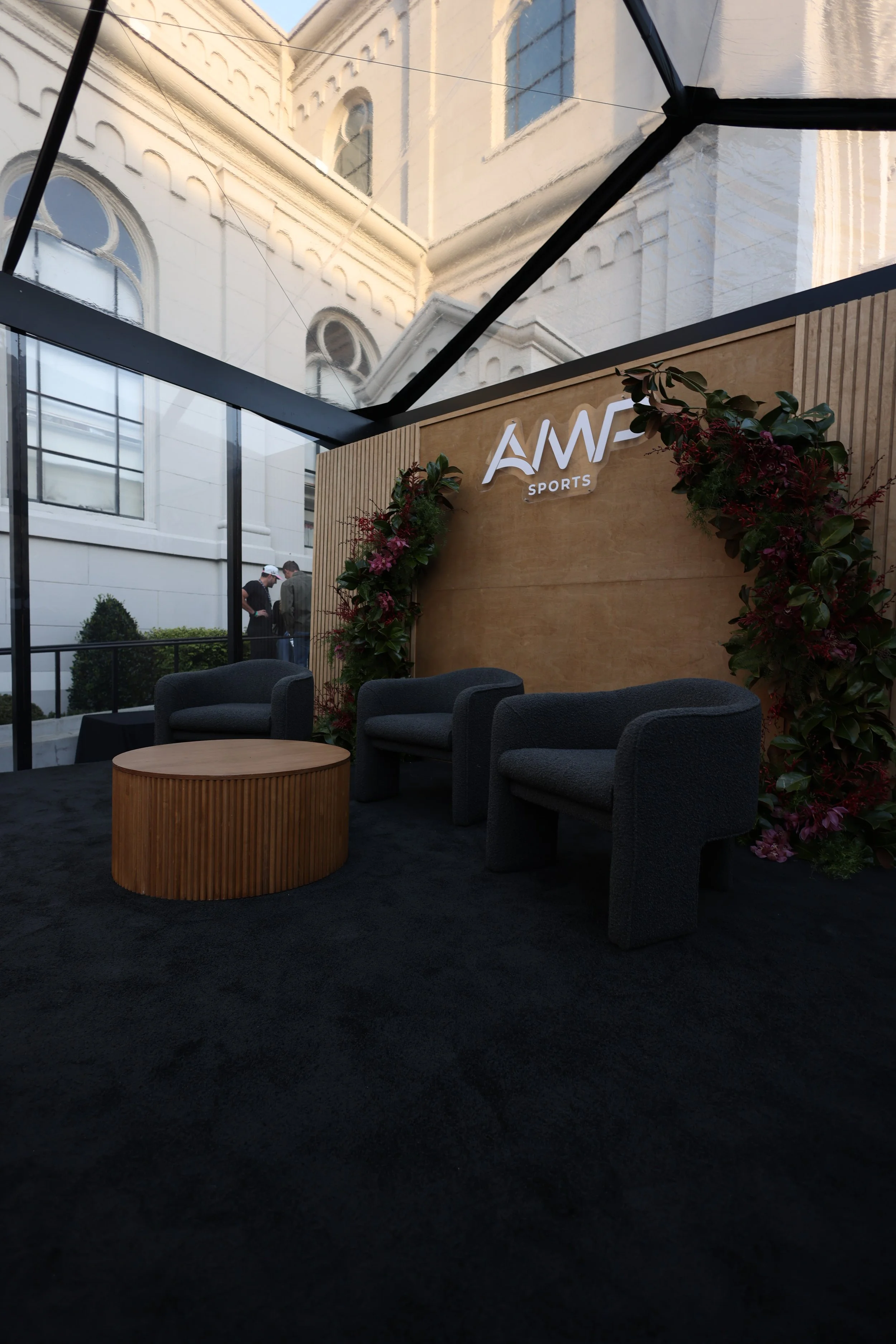Interior view of a modern enclosed patio or lounge area with black carpet flooring, black chairs, a round wooden table, a wooden wall with flowers and the sign 'AVE Sports', glass walls, and reflections of an ornate white building with arched windows