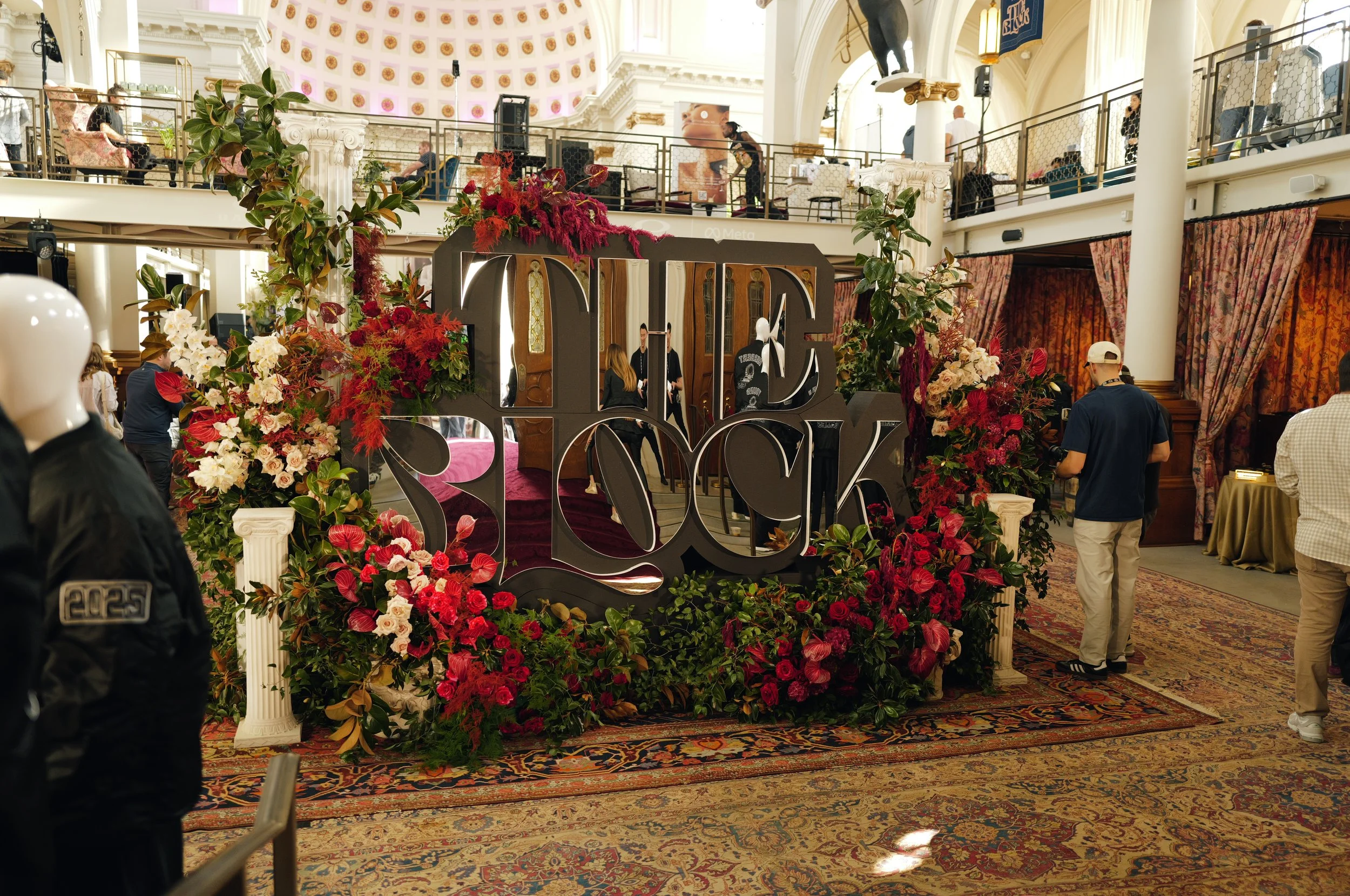 Interior of a hotel lobby or event space decorated with a large floral arrangement, a 3D love sign, and people gathering and walking around. The space features high ceilings, ornate architectural details, a second-floor balcony, and an ornate carpet.