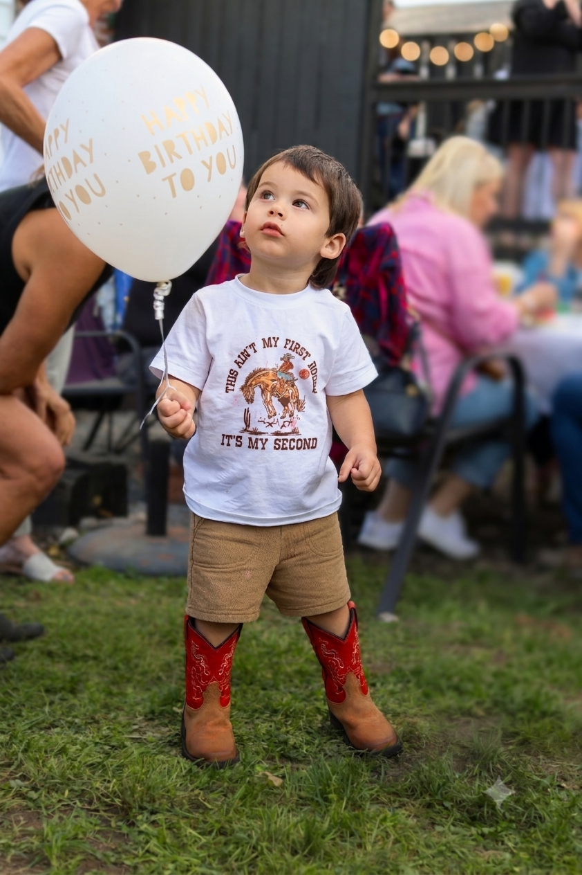 A young boy holding a white balloon with gold writing that says "Happy Birthday to You" at an outdoor birthday celebration. He wears a white t-shirt with a graphic of a goat and the text "This ain't my first rodeo, it's my second," brown shorts, and 