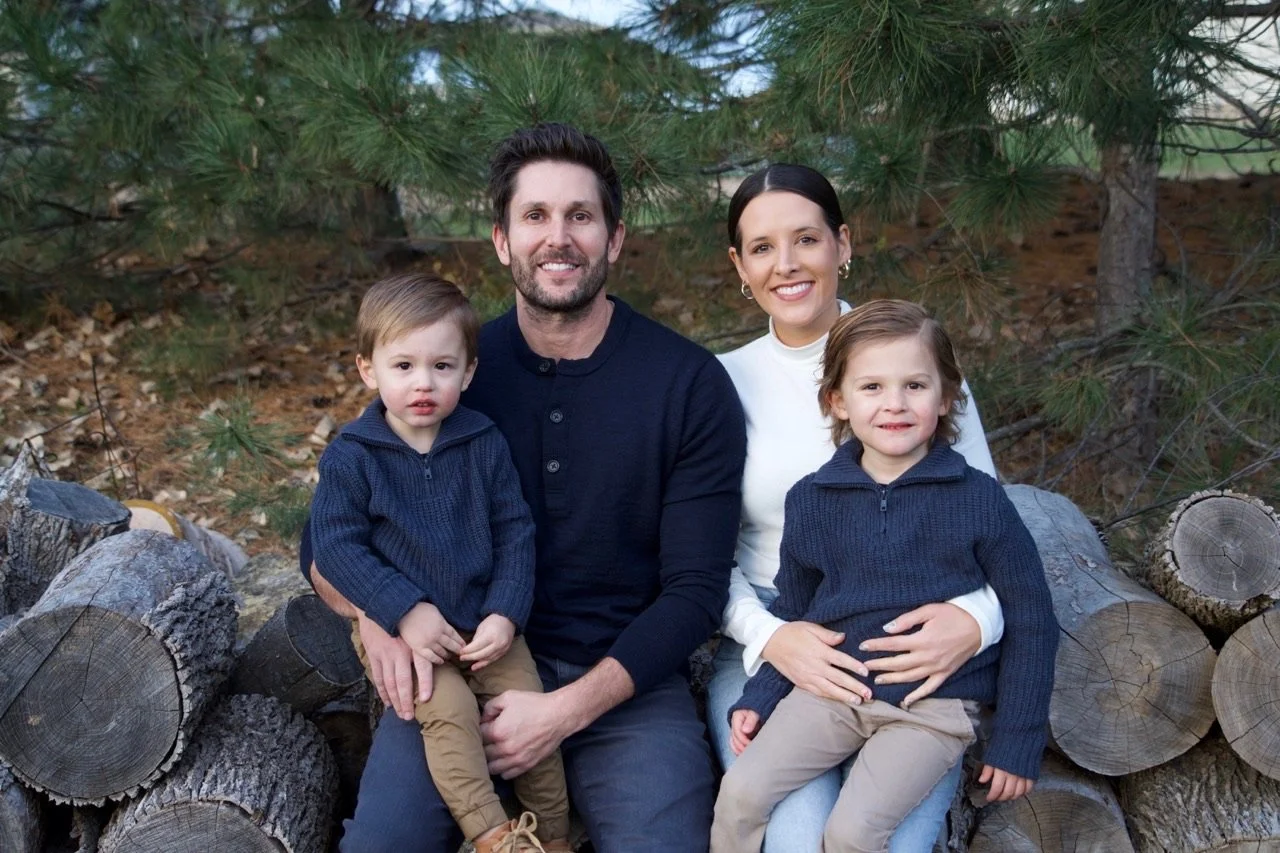 A family of four sitting outdoors on logs near a pine tree, with two young boys and their parents smiling at the camera.
