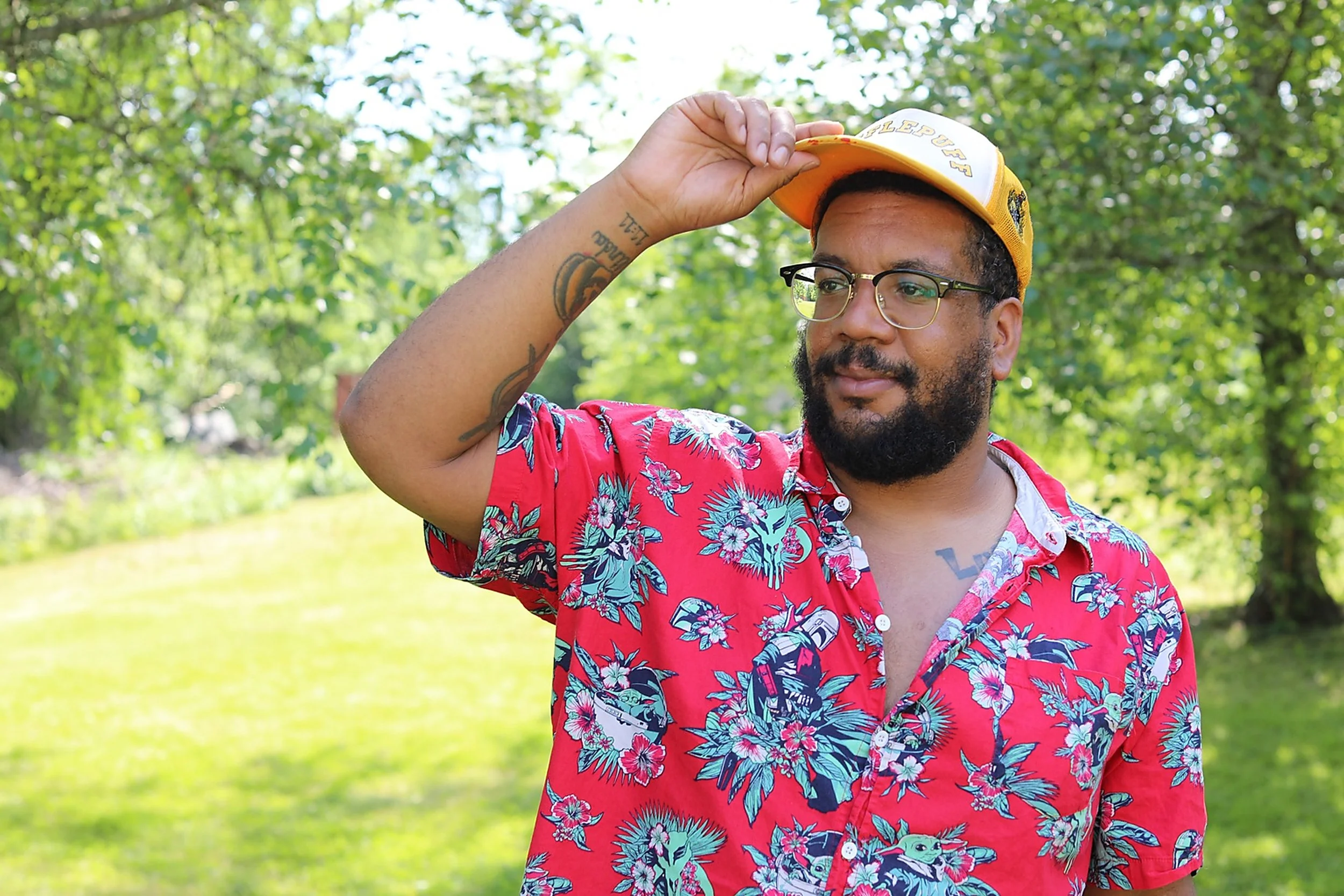 A man with glasses and a beard is outdoors, wearing a red Hawaiian shirt with a floral pattern and a yellow cap, adjusting the cap with his right hand. The background shows green trees and a bright sky.