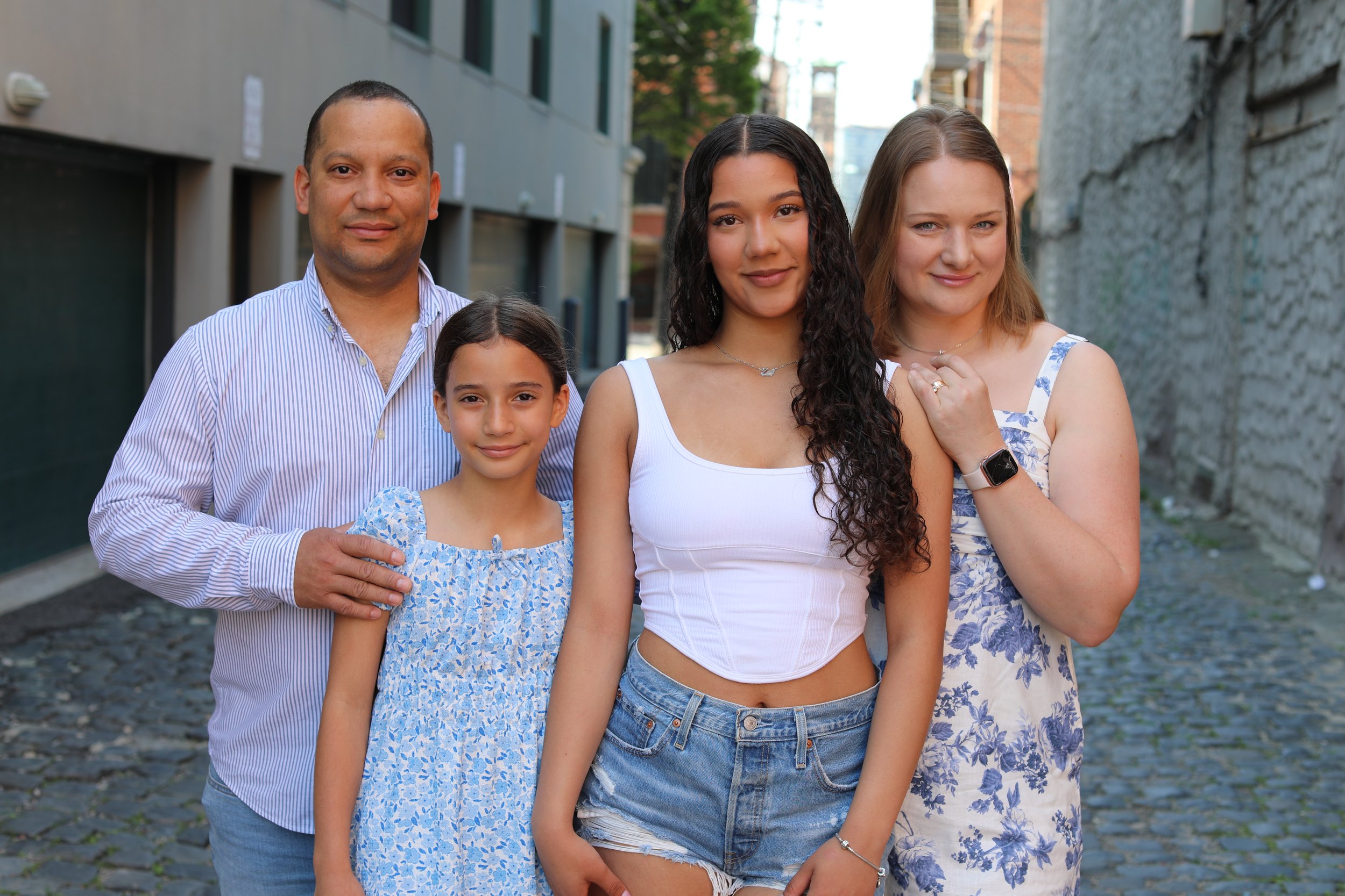 A diverse group of five people, including three women and two children, standing outdoors on a cobblestone street between buildings, smiling at the camera.