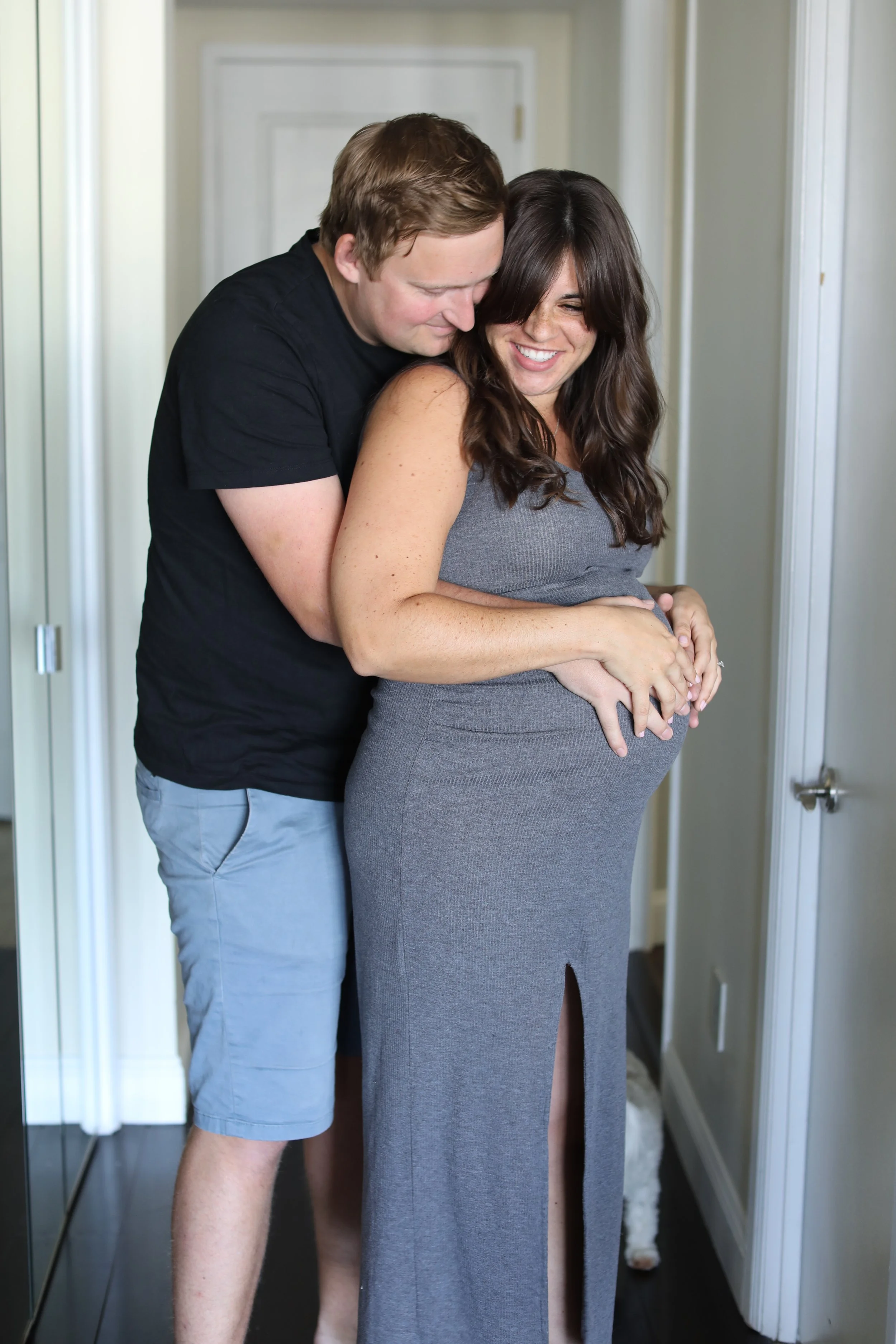 A man and a pregnant woman are hugging each other and smiling in a hallway, with a dog in the background.