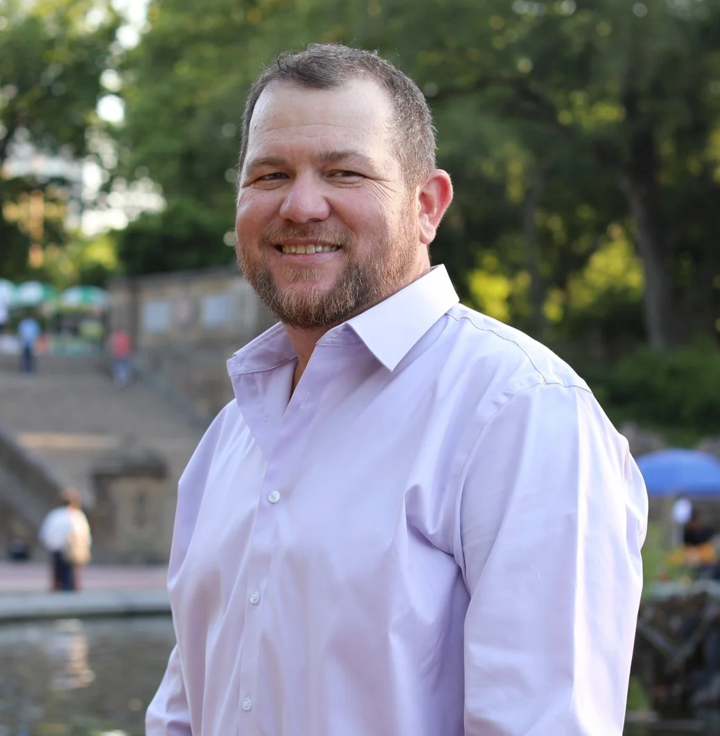 A smiling man with short dark hair and a beard, wearing a white dress shirt, standing outdoors with trees and some people in the background during daytime.
