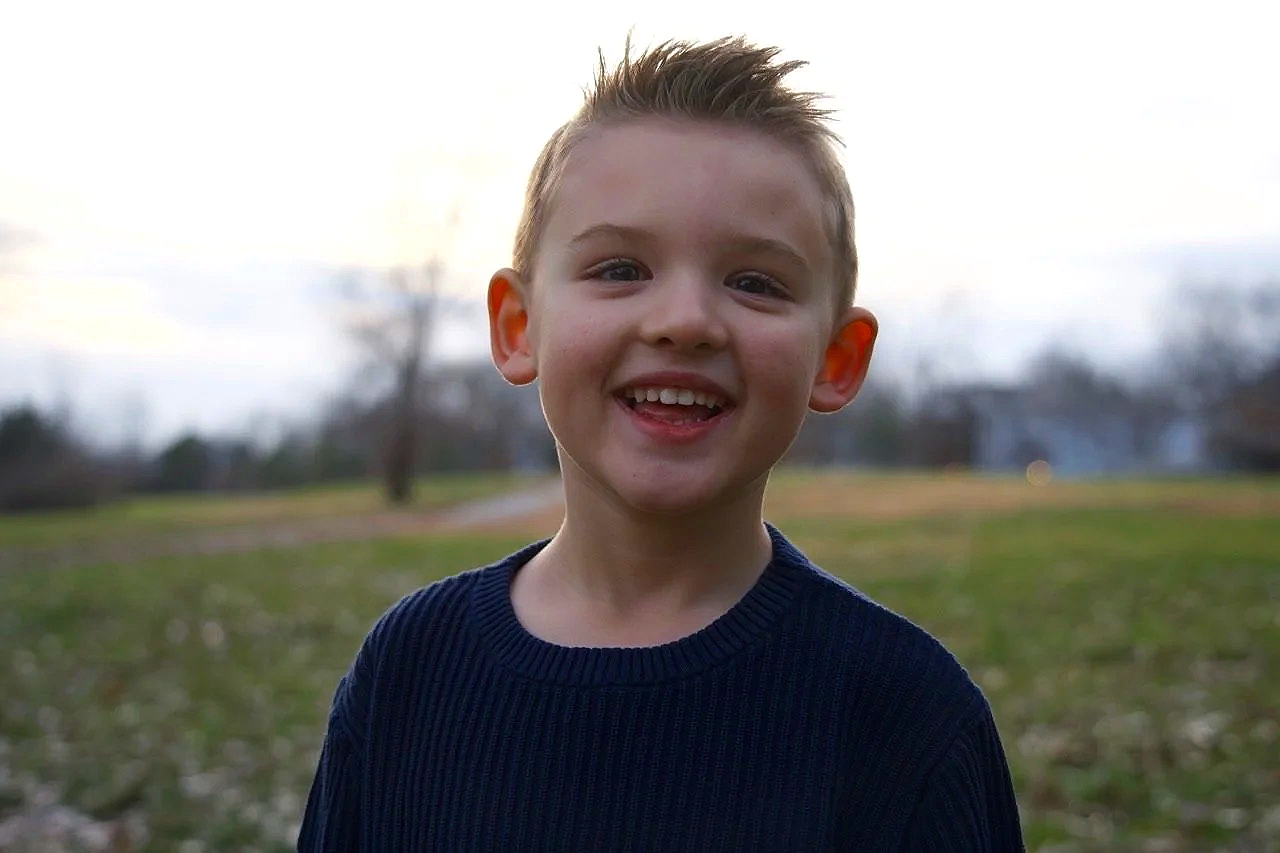 A young boy with short, spiked hair smiling outdoors in a park during daytime with trees and grass in the background.