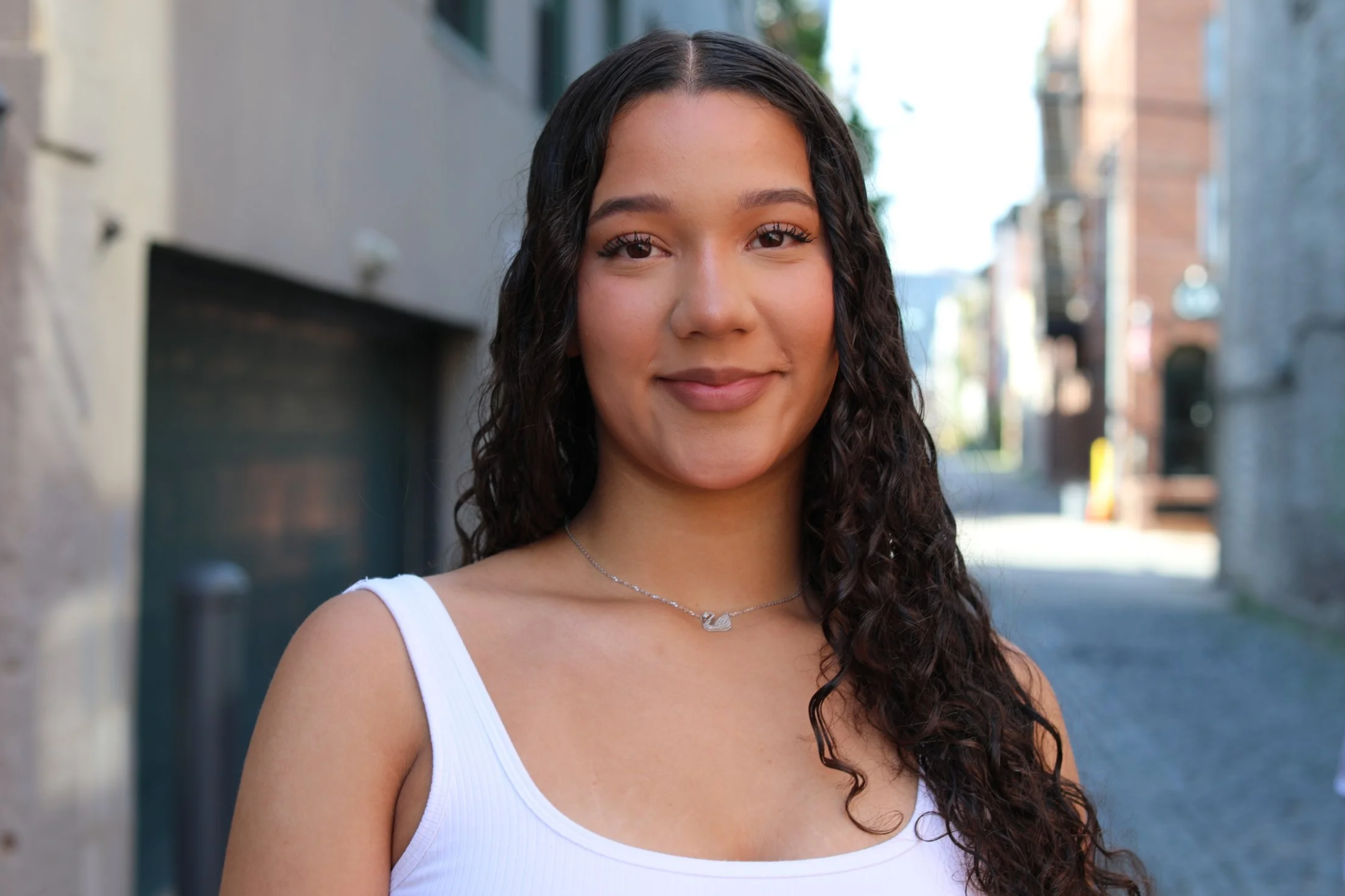A young woman with long wavy dark hair, wearing a white sleeveless top and a delicate necklace, standing outdoors in an urban setting with buildings and a narrow alley in the background.