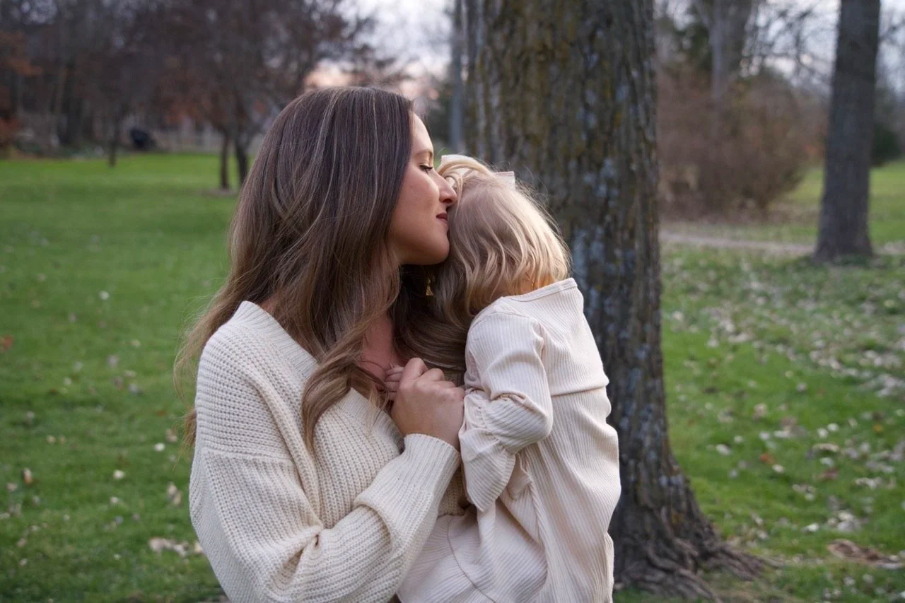 A woman with long brown hair and a white sweater is holding and comforting a young girl with blond hair and a cream-colored outfit in a park with grass and trees.