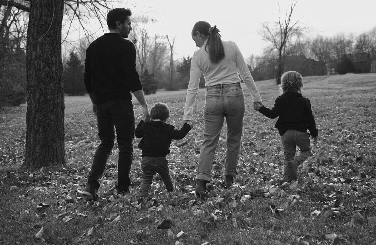 A black and white photo of a family walking outdoors in a park with trees and fallen leaves. The group is holding hands and walking away from the camera.