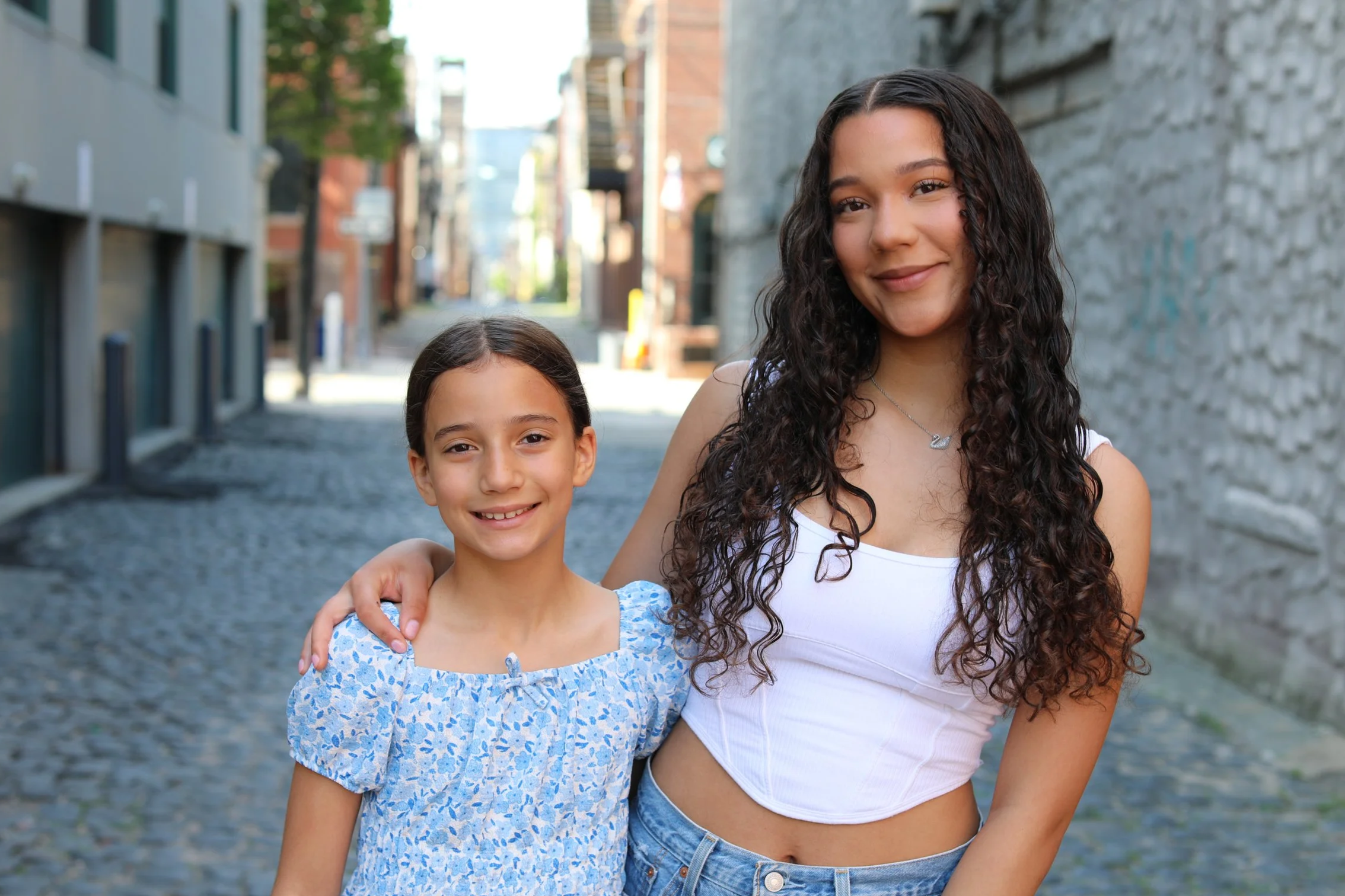 A young girl and woman with curly dark hair smiling outdoors on a city street, with the woman’s arm around the girl.
