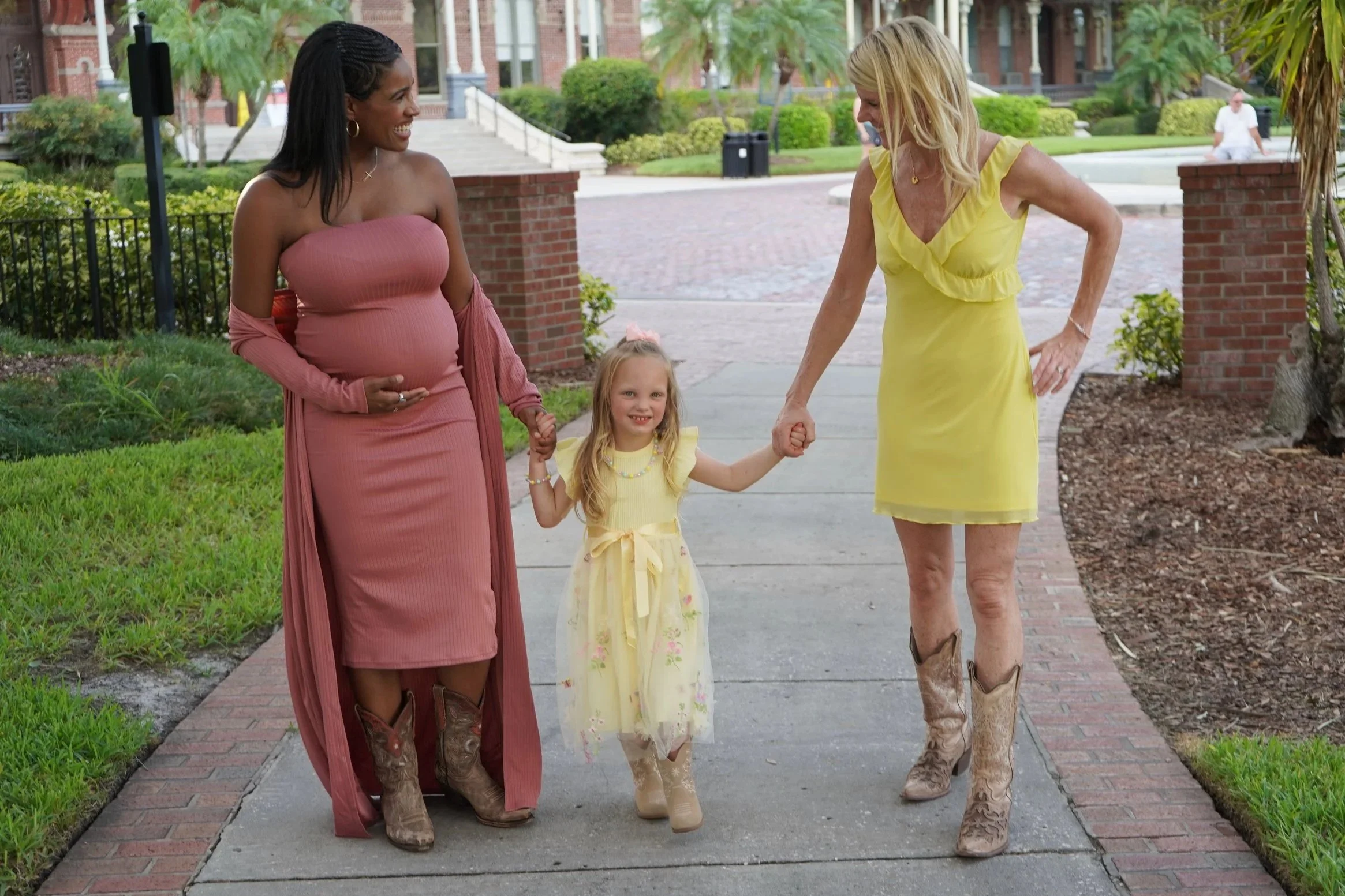 Three women and a young girl holding hands walking outdoors on a sidewalk. The women are dressed in colorful dresses and cowboy boots, and the girl is wearing a yellow dress with floral accents. The background includes green grass, bushes, trees, and