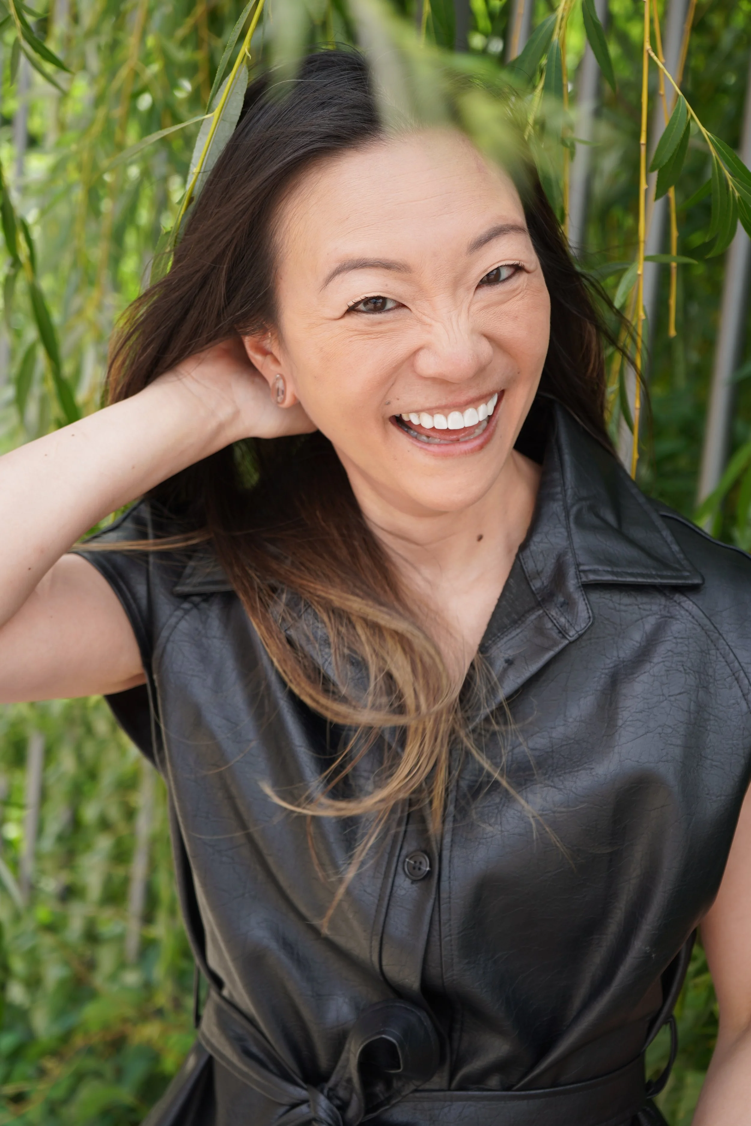 A woman with long brown hair smiling and wearing a black leather vest outdoors near green plants.