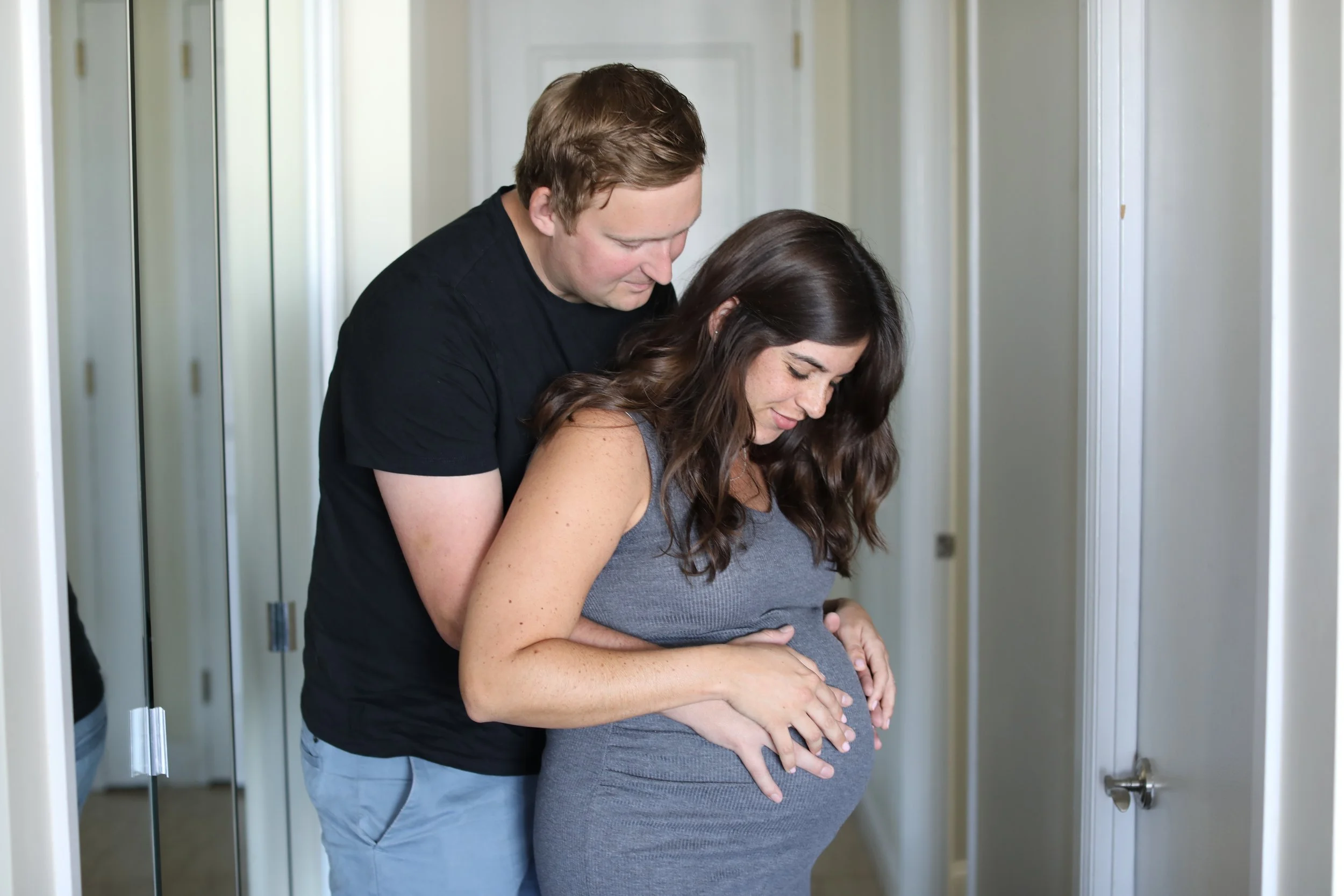 A man and woman, with the man standing behind the woman, both leaning forward, with their hands on her pregnant belly, in a hallway with mirrored closet doors.