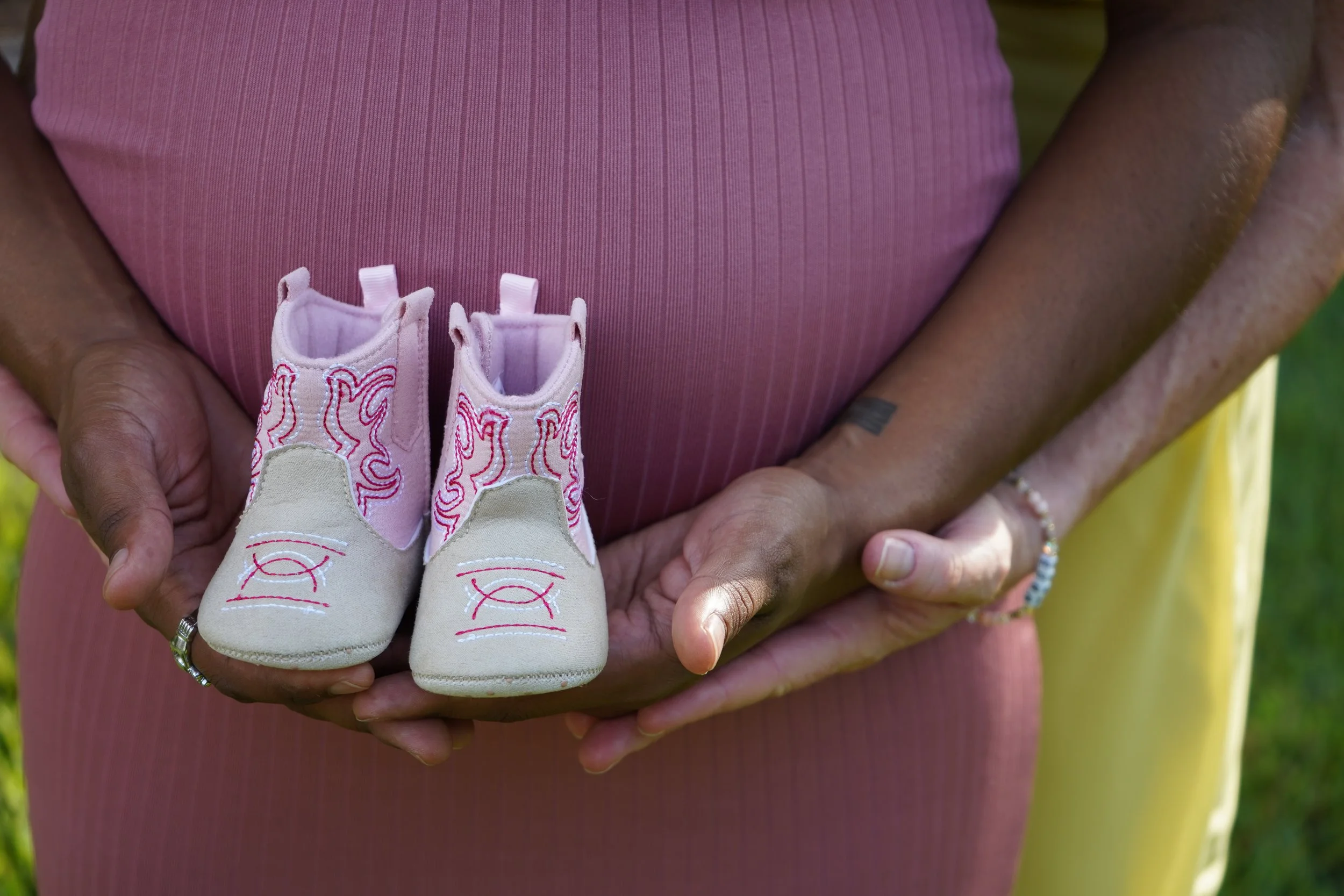 Person holding a pair of tiny beige sneakers with pink stitching in front of their chest, wearing a pink top, with hands visible, one with a ring and bracelet, and a small tattoo on their wrist.