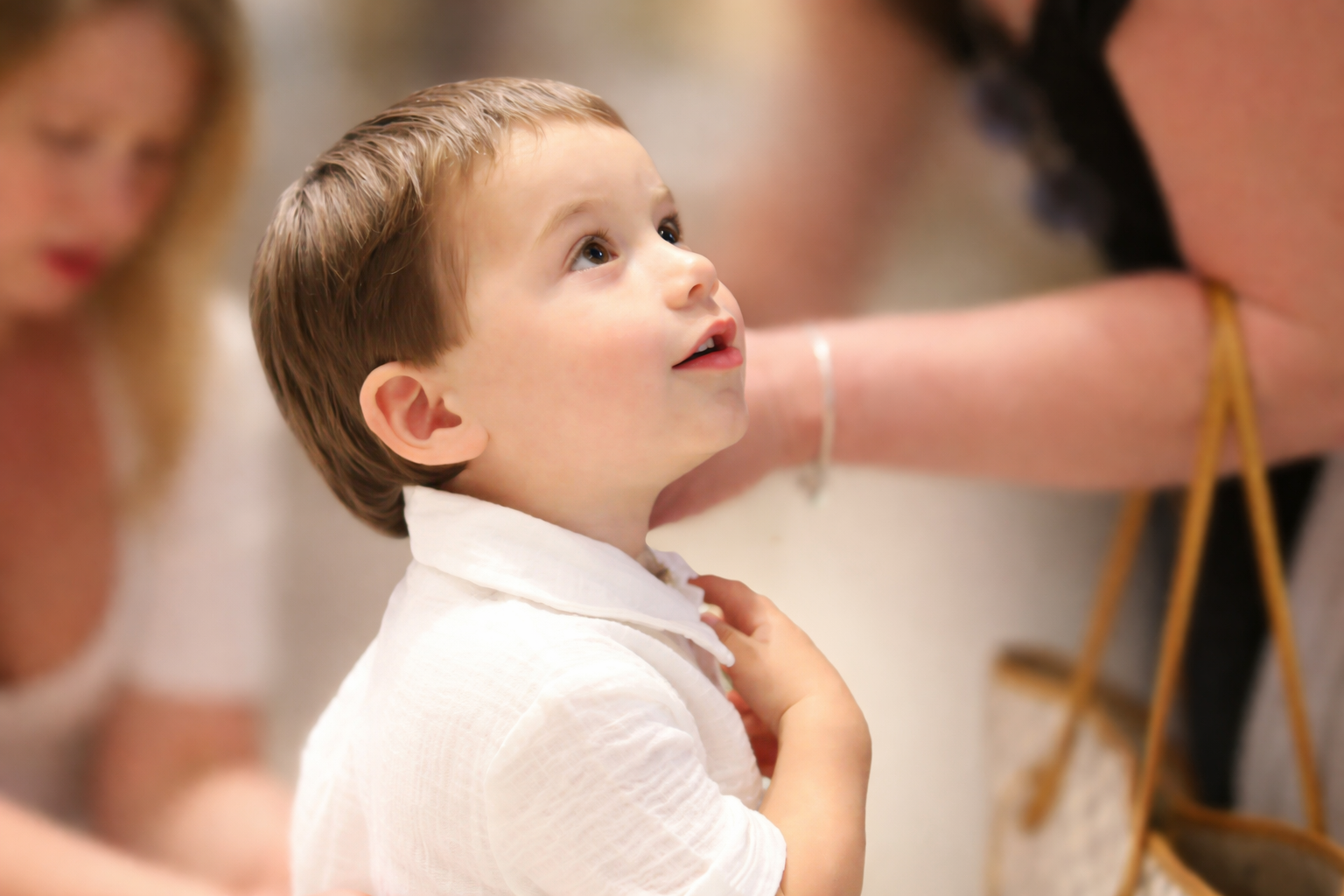 A young boy with light brown hair and fair skin looks up with wonder, wearing a white shirt. An adult woman with long blond hair is partially visible on the left, and another woman with dark hair and a bracelet is partially visible on the right, both