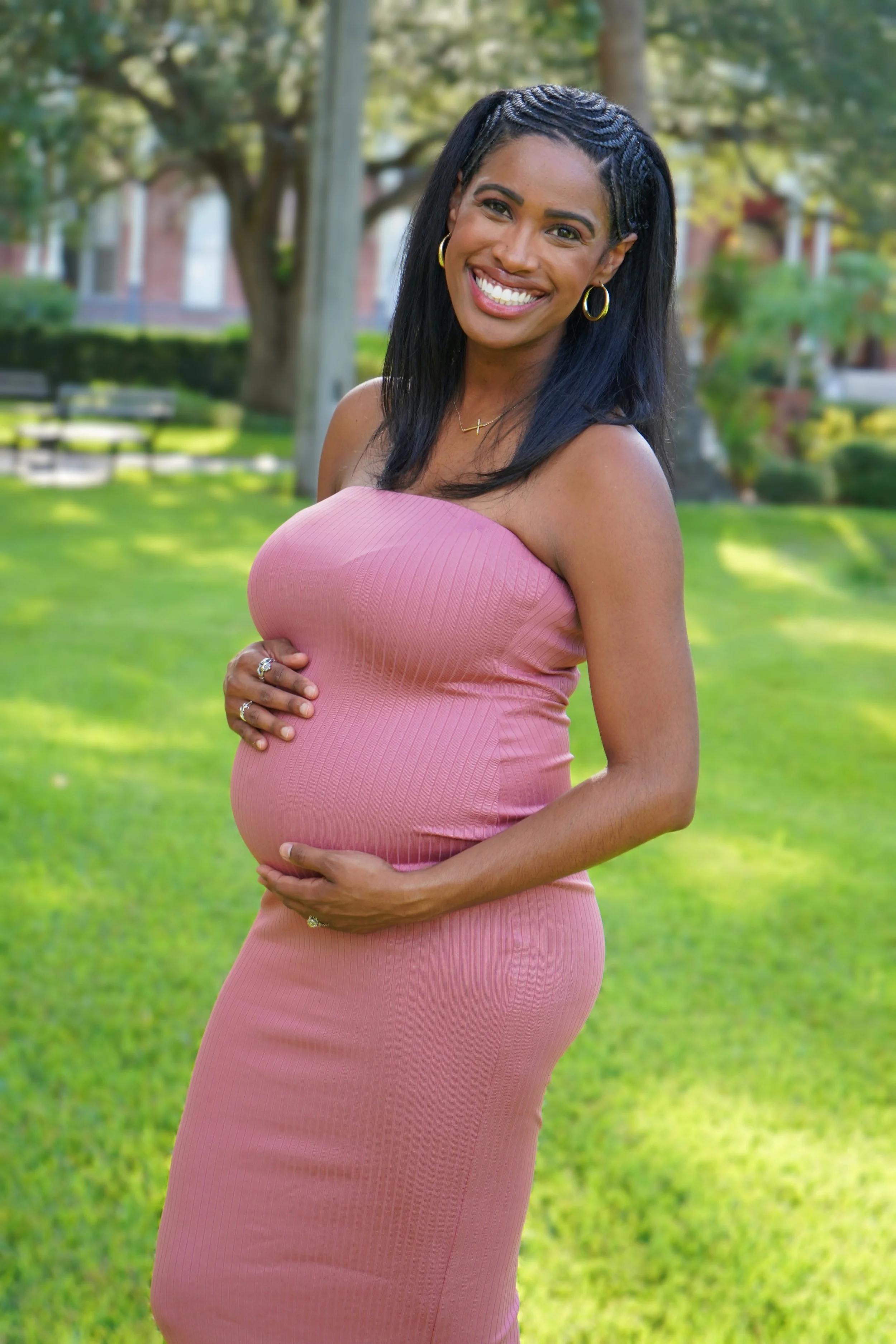 A smiling pregnant woman with dark hair, wearing gold hoop earrings, a delicate cross necklace, and a pink strapless dress, standing outdoors in a park with green grass, trees, and benches in the background.
