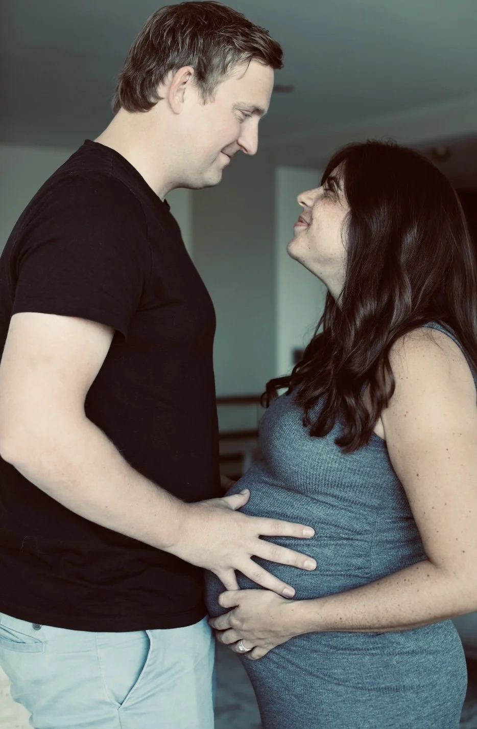 A pregnant woman and a man standing close, looking into each other's eyes lovingly, with the man's hand on her belly.