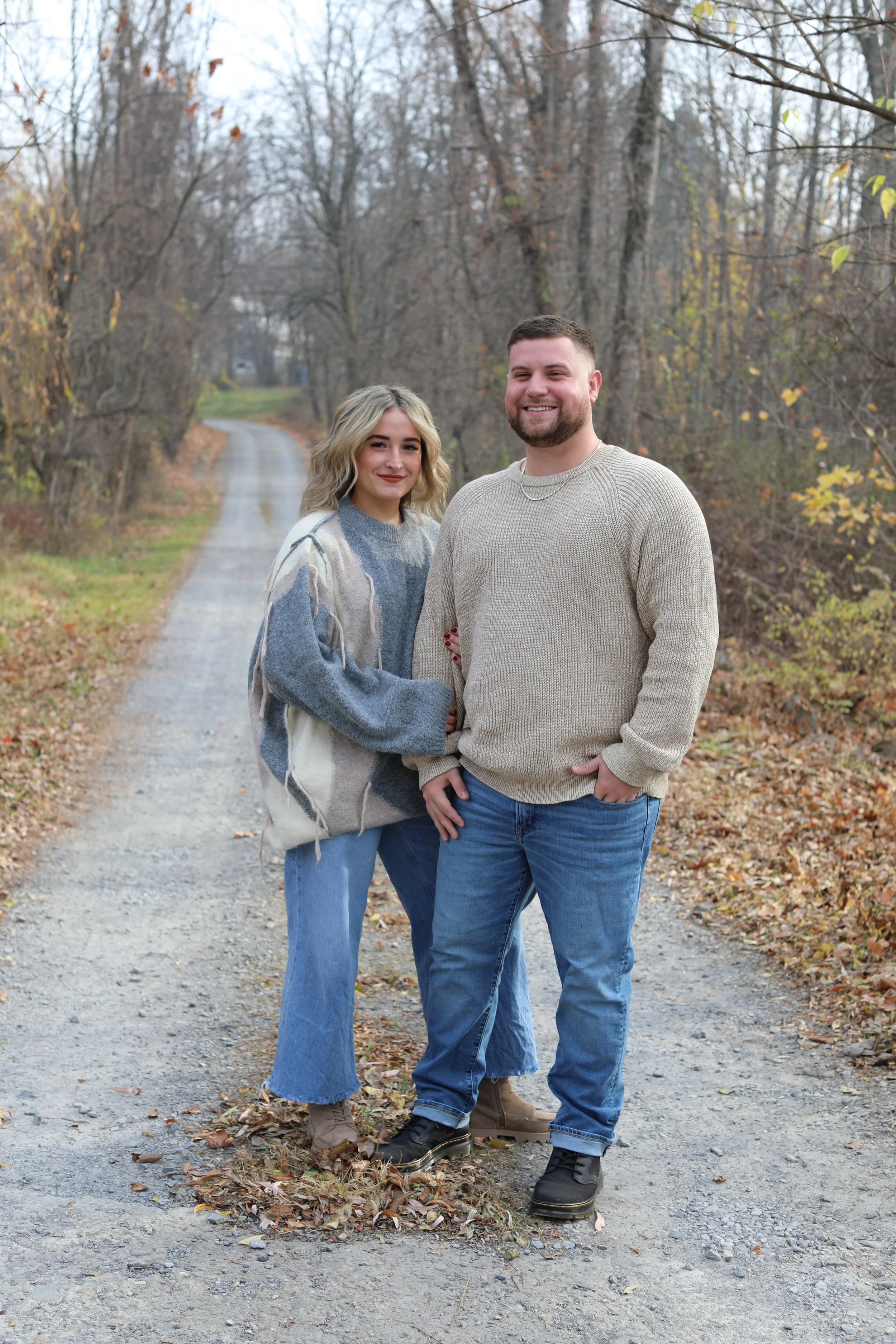 A happy couple standing on a dirt road in a wooded area during fall, with leafless trees and scattered leaves on the ground.