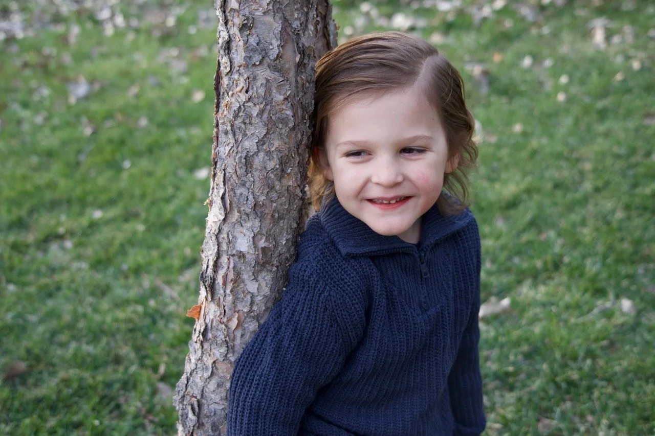 A young girl with brown hair and a navy blue sweater leaning against a tree outdoors, smiling softly.