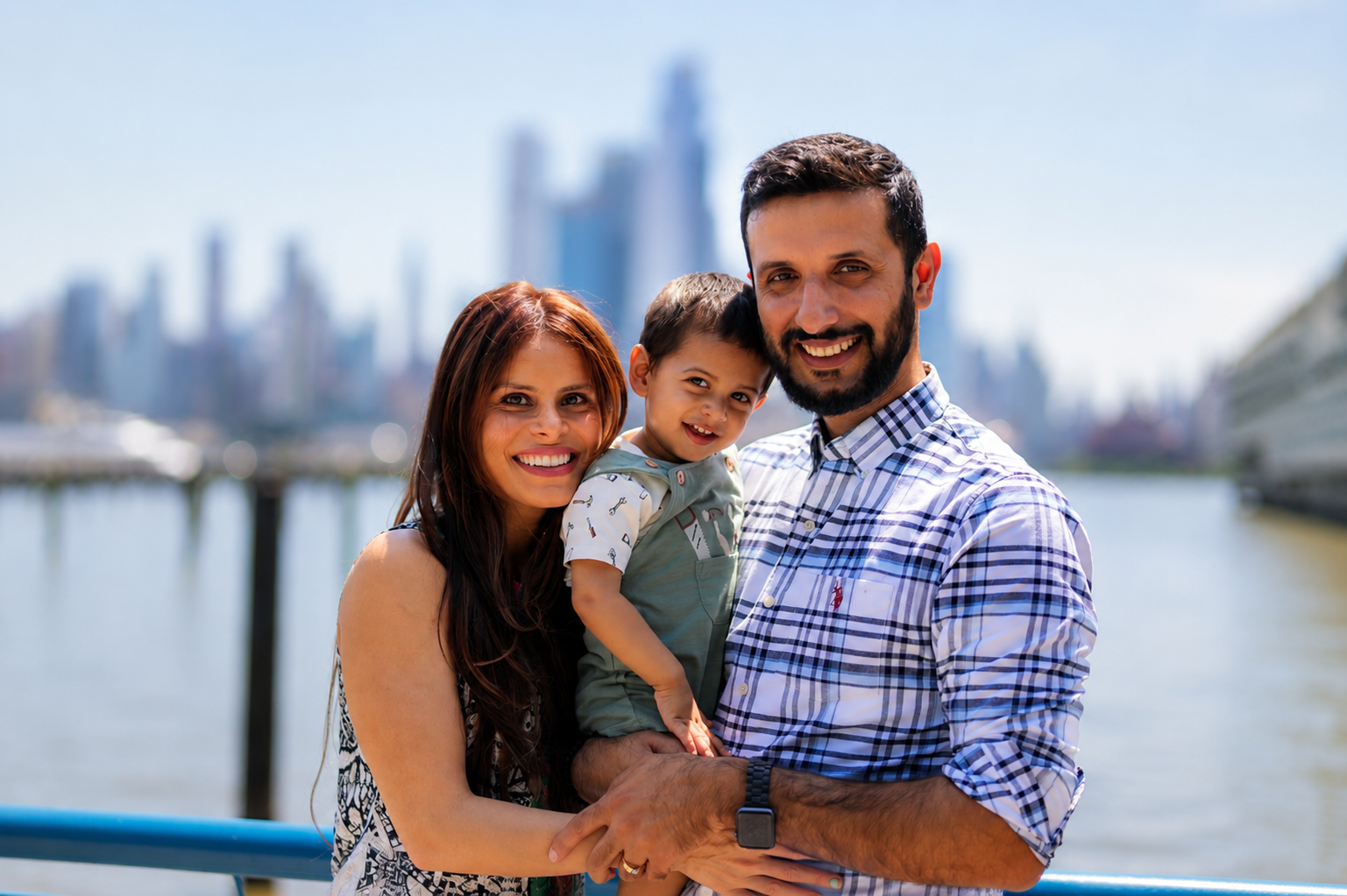 A happy family of three, a woman, a man, and a young boy, smiling outdoors by a body of water with a city skyline in the background.
