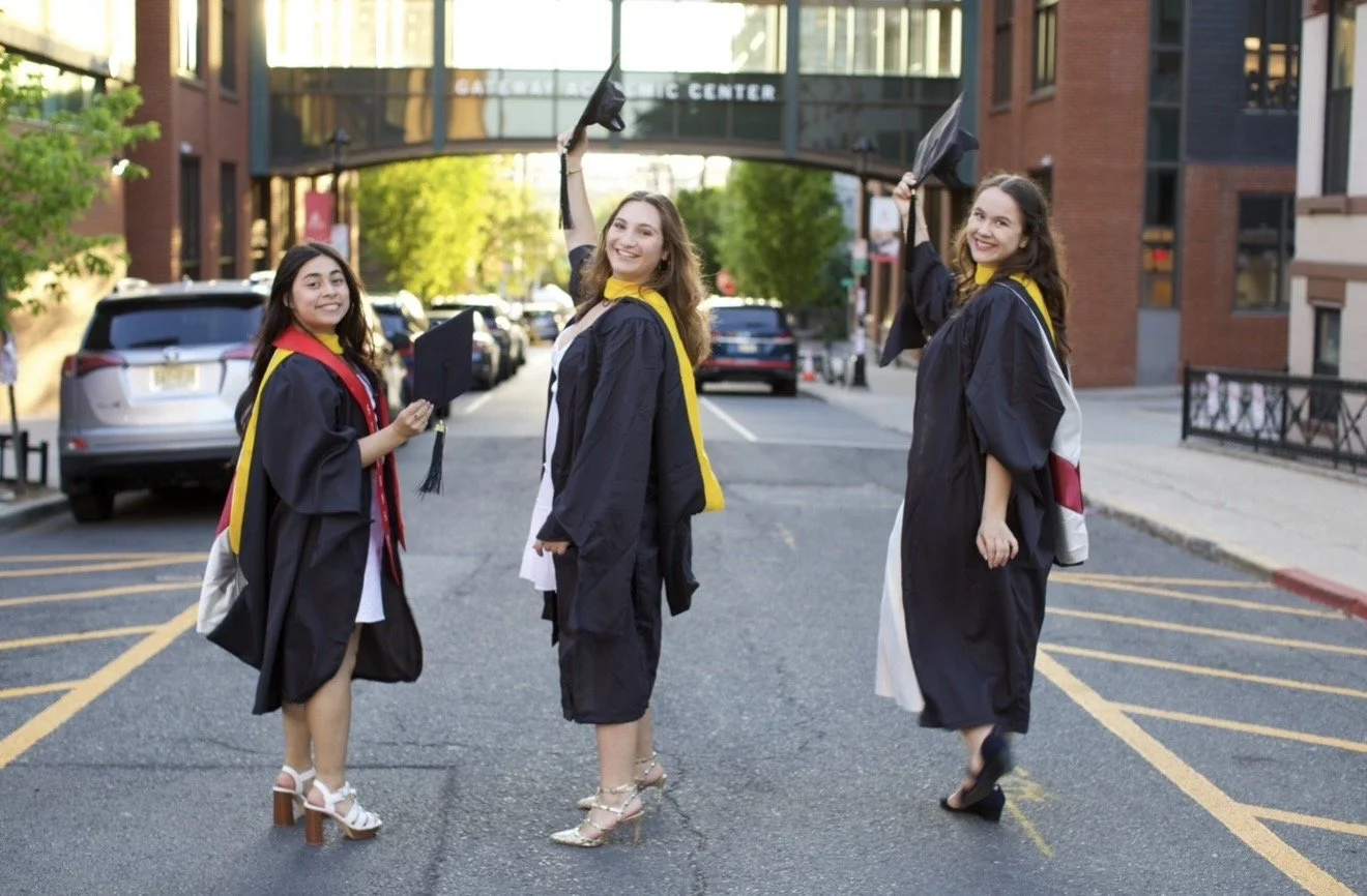 Three women in caps and gowns celebrating graduation outdoors on a city street.
