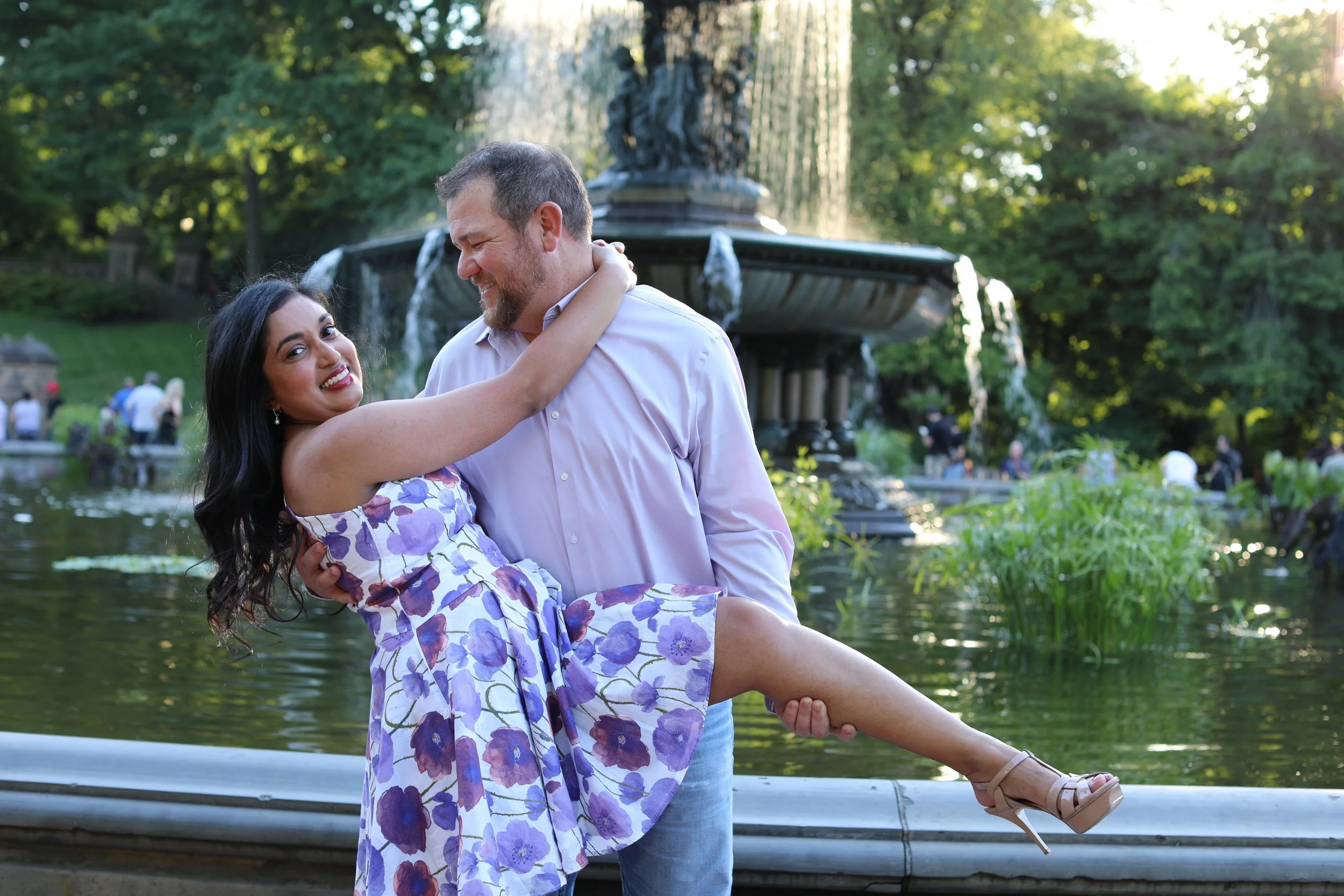 A couple enjoying a romantic moment by a fountain in a park during daytime. The woman is sitting on the man's lap with her arms around his neck, smiling, while the man is holding her with a smile. She is wearing a purple floral dress and heels, and h