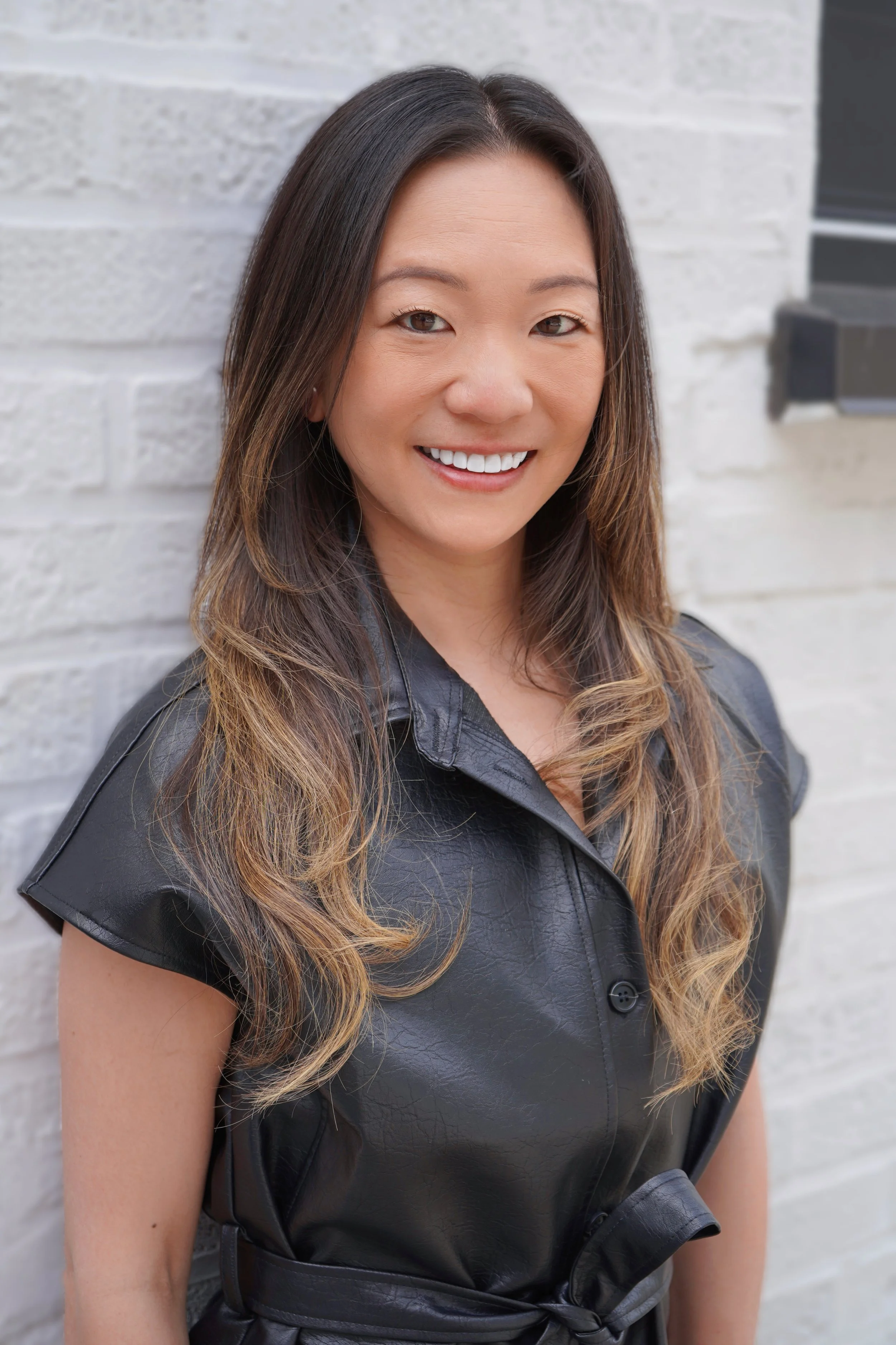 Portrait of a smiling woman with long wavy hair, wearing a black leather dress, standing against a white brick wall.