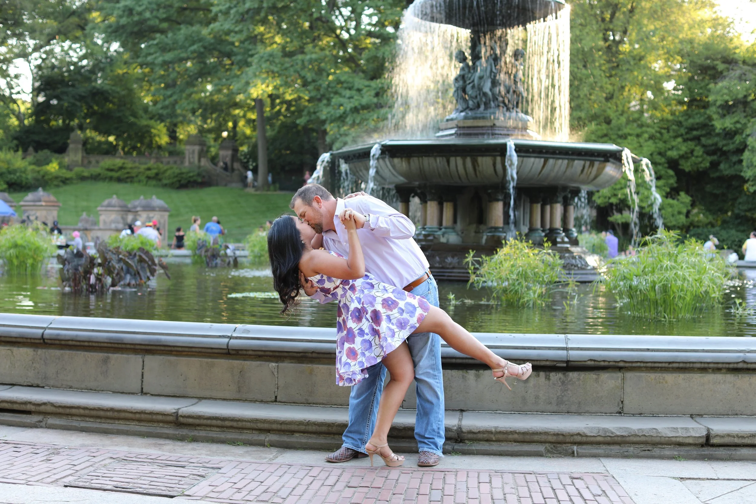 A man and woman sharing a romantic kiss by a fountain in a park with green trees and people in the background.