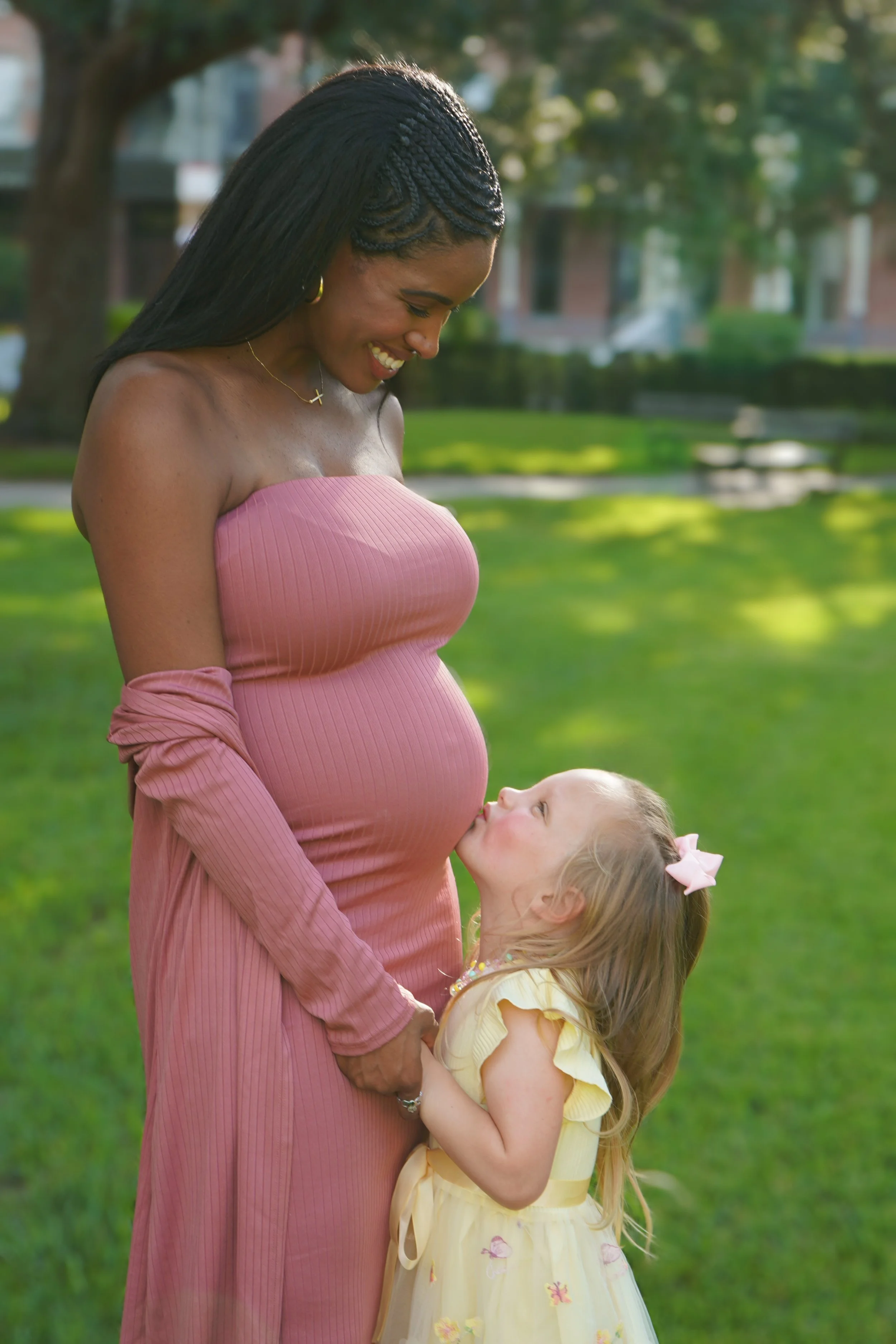 A woman with braids and a pink dress looks down and smiles at a young girl with a yellow dress and a pink bow in her hair, who is kissing her pregnant belly. They are outdoors in a park with green grass and trees.