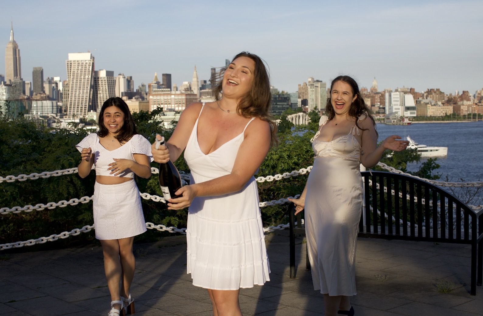 Three women in white dresses celebrating outdoors with city skyline and water in the background. One woman opens a bottle of champagne, and others are smiling and laughing.