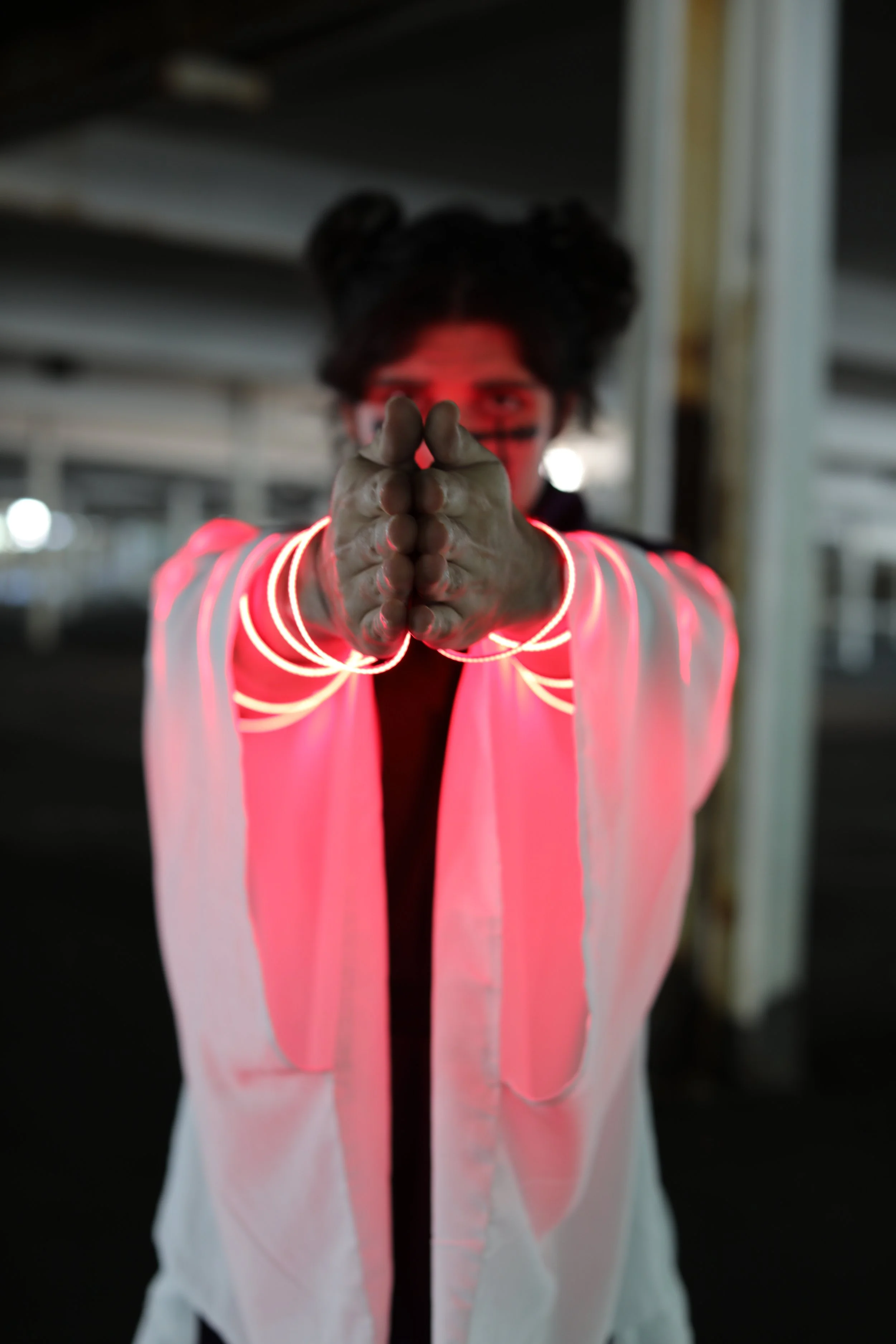 Person with dark hair styled in buns, wearing a white jacket with red neon lights on the sleeves, and holding their hands together in front of their face in a dimly lit environment.