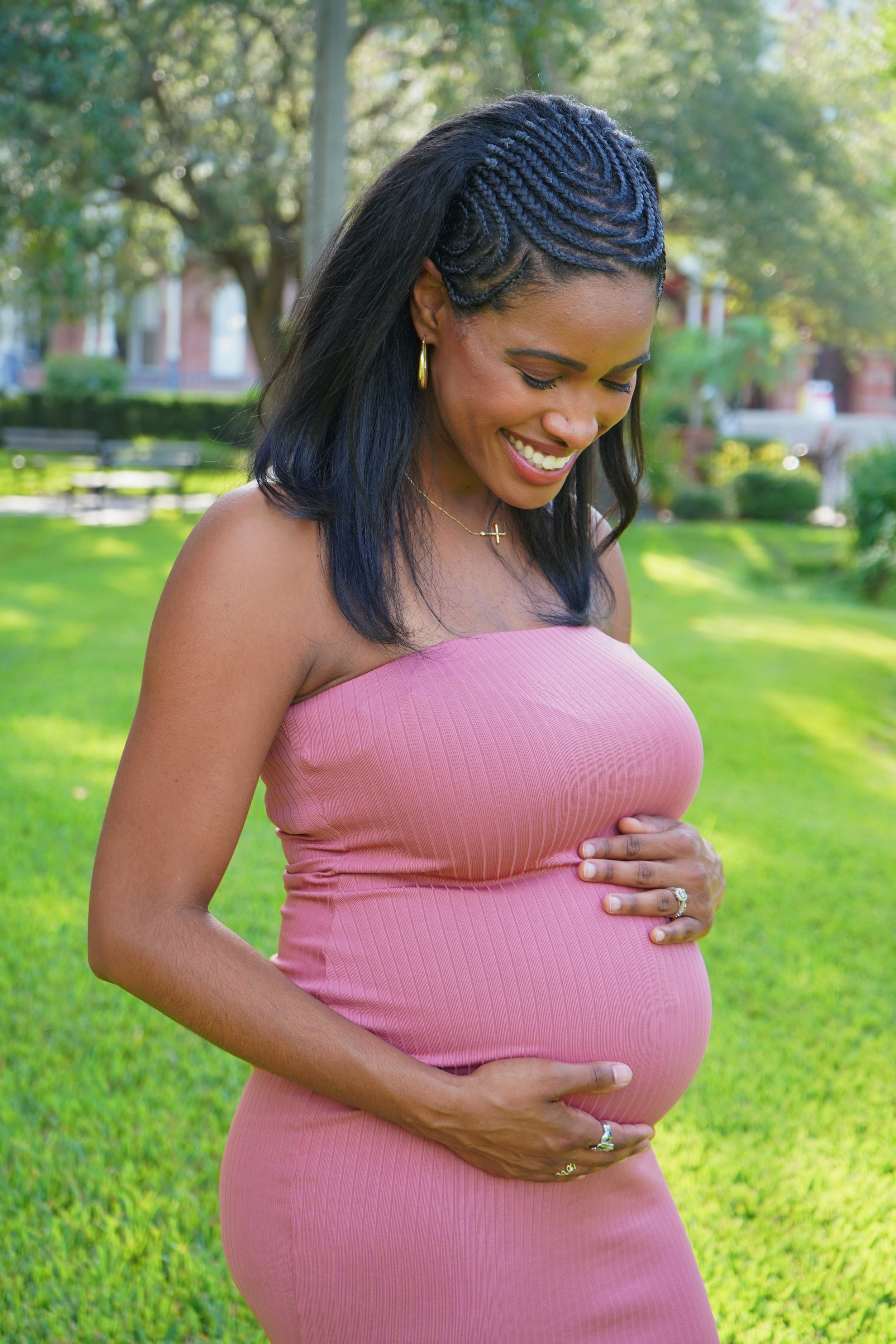 A pregnant woman with dark hair styled in blue braids, wearing a pink strapless dress, standing outdoors on a sunny day with green trees and grass, smiling and looking down at her baby bump.