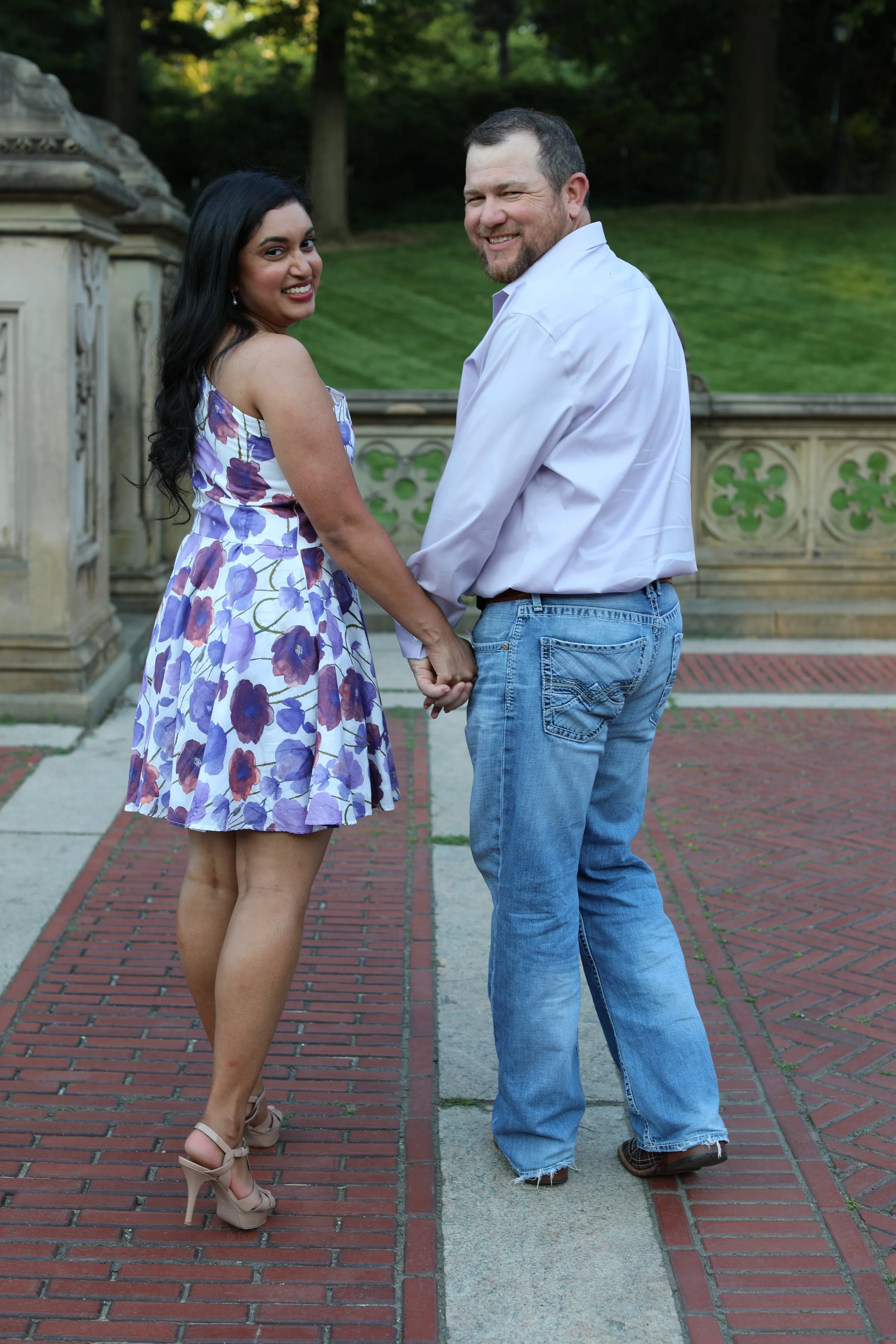 A couple holding hands and smiling outdoors on a brick path with greenery and trees in the background.