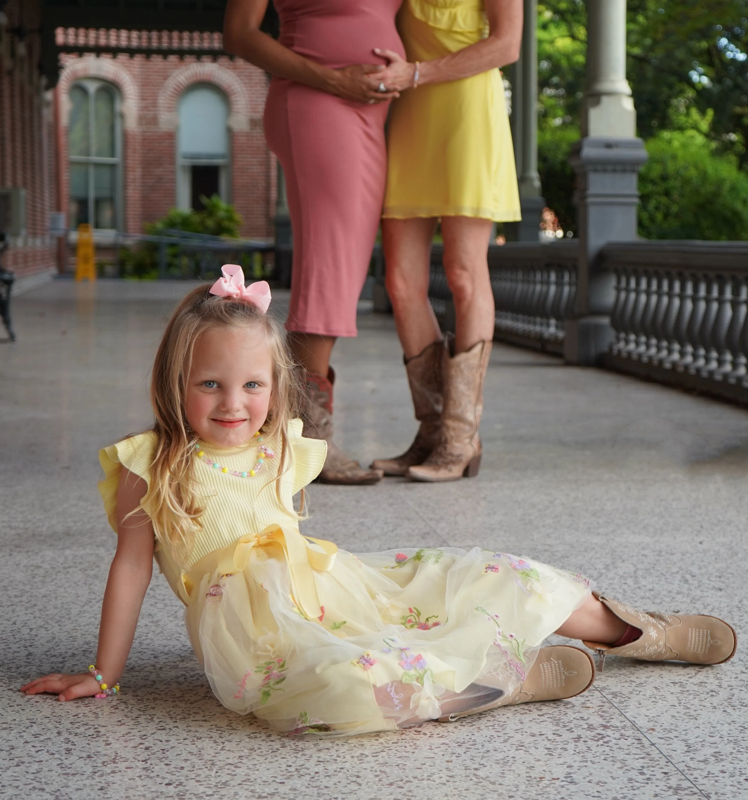 A young girl with blonde hair and blue eyes, wearing a yellow dress with a pink bow in her hair, sitting on the floor with her legs crossed and smiling at the camera. There are adults standing behind her, one in a pink outfit and the other in a yello