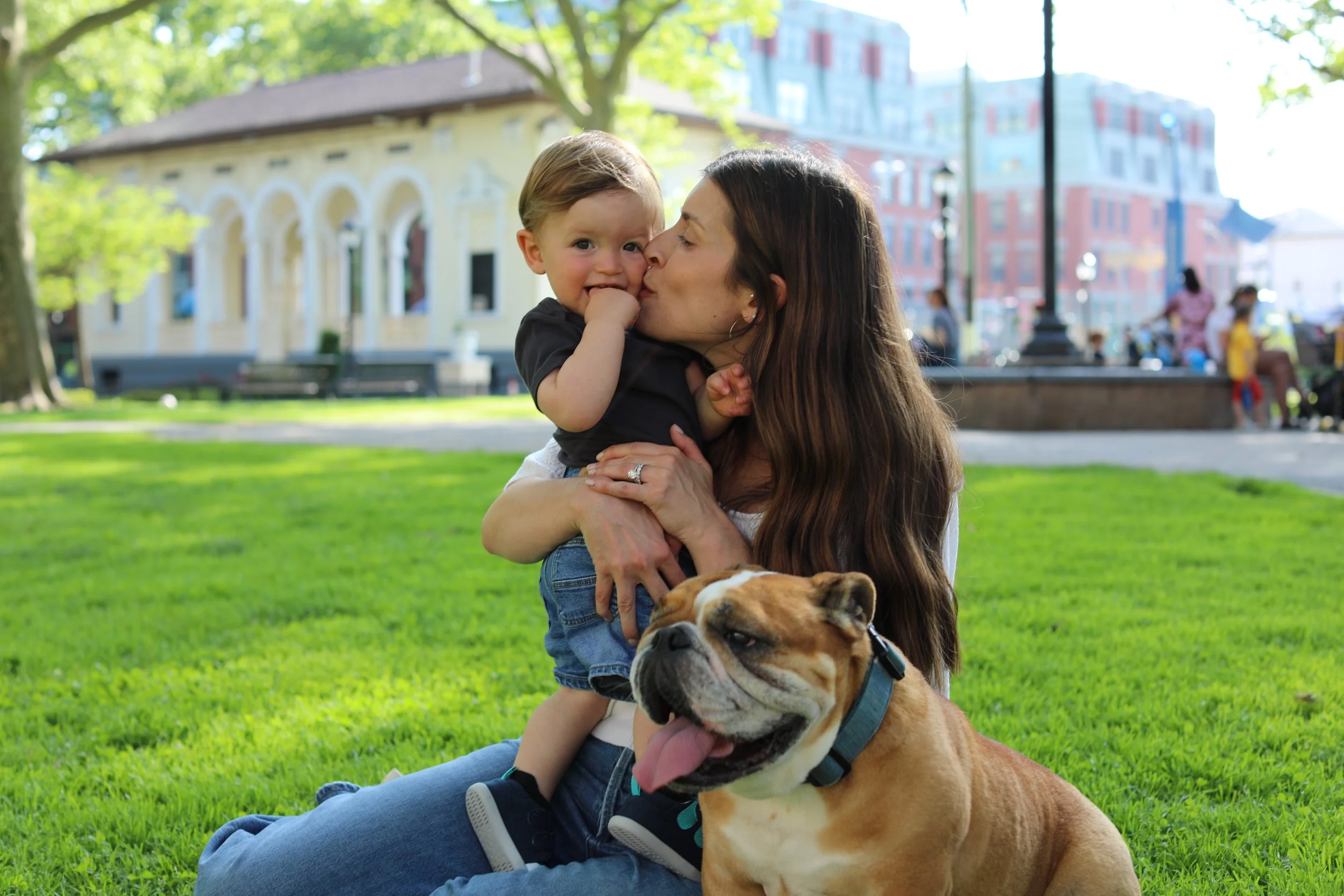 A woman and young boy sharing a kiss outdoors in a park, with a bulldog sitting on the grass in front of them. The boy has his hand near his mouth, and the scene has green grass, trees, and buildings in the background.